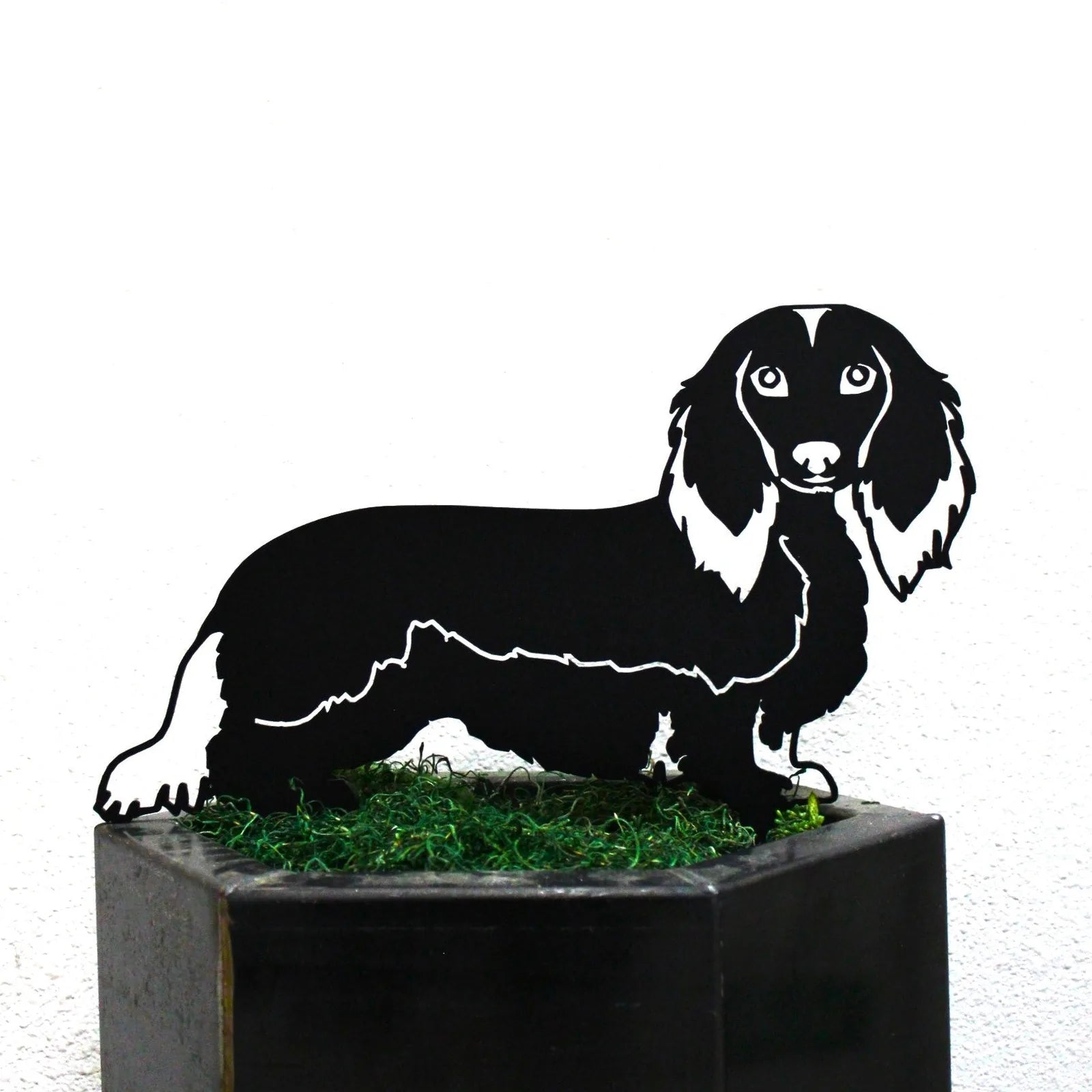 Black metal silhouette of a longhaired Dachshund with detailed features, resting on green decorative grass.