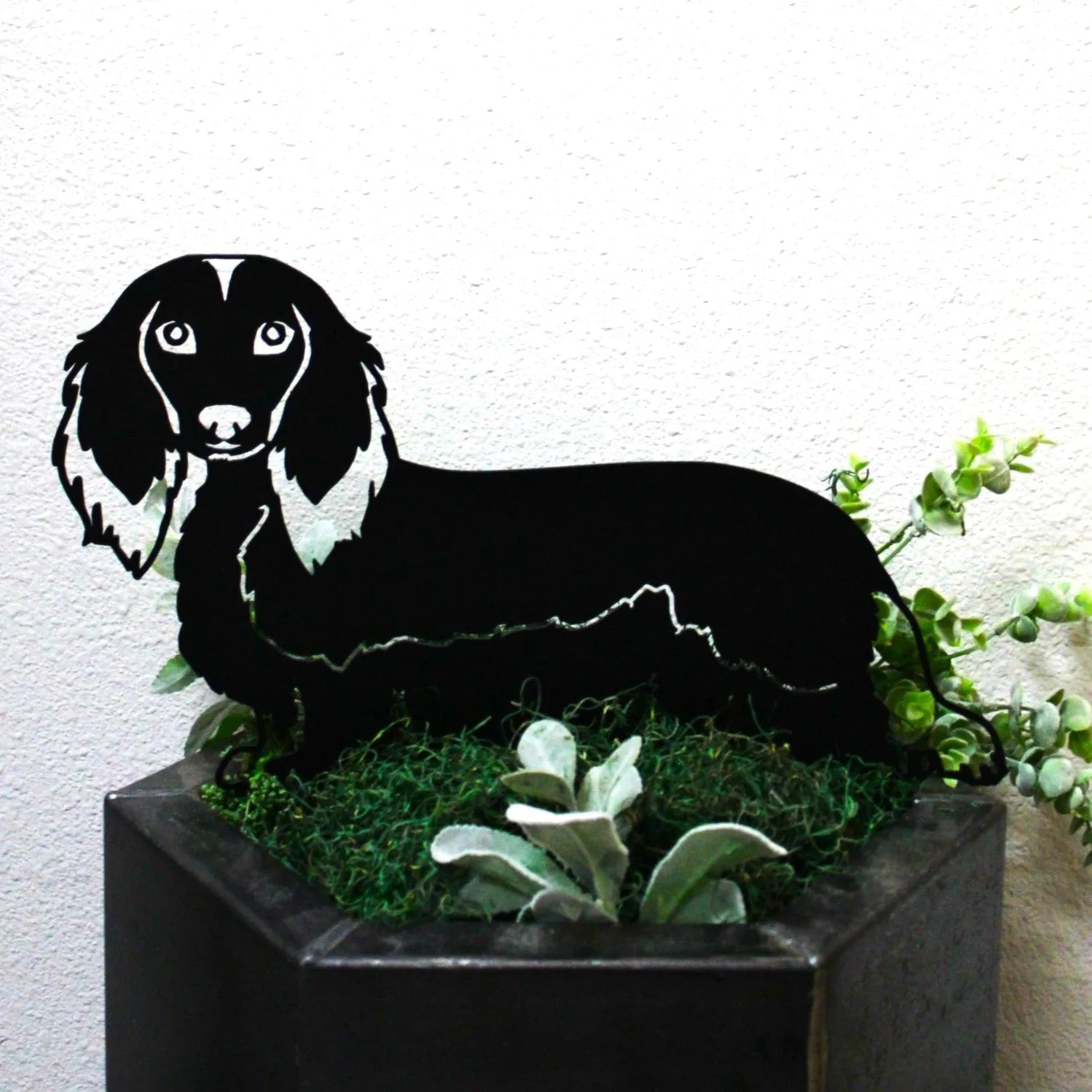 Black metal silhouette of a longhaired Dachshund, displayed among greenery in a planter.