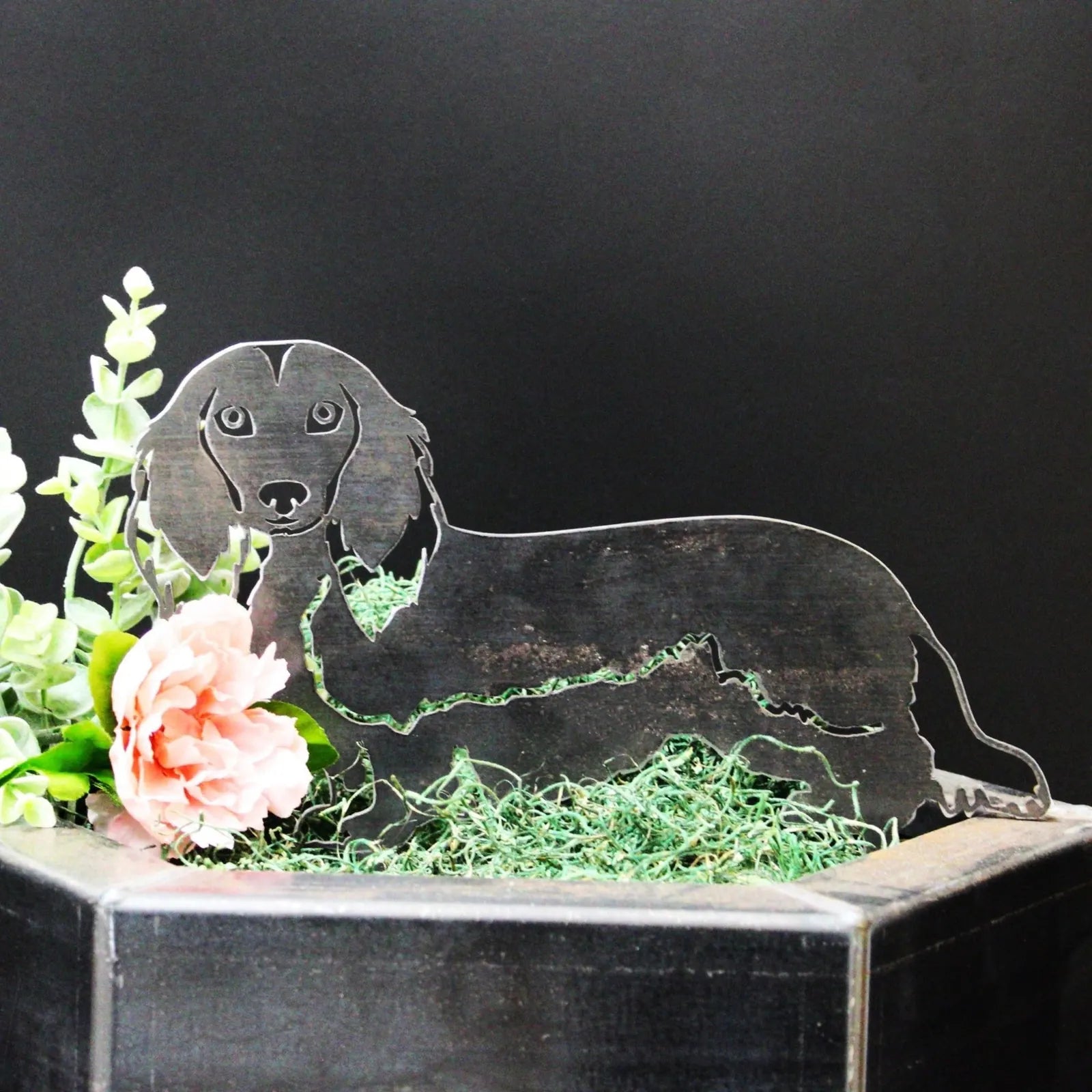 Handmade metal silhouette of a longhaired Dachshund in Corten steel, surrounded by pink flowers and green moss.