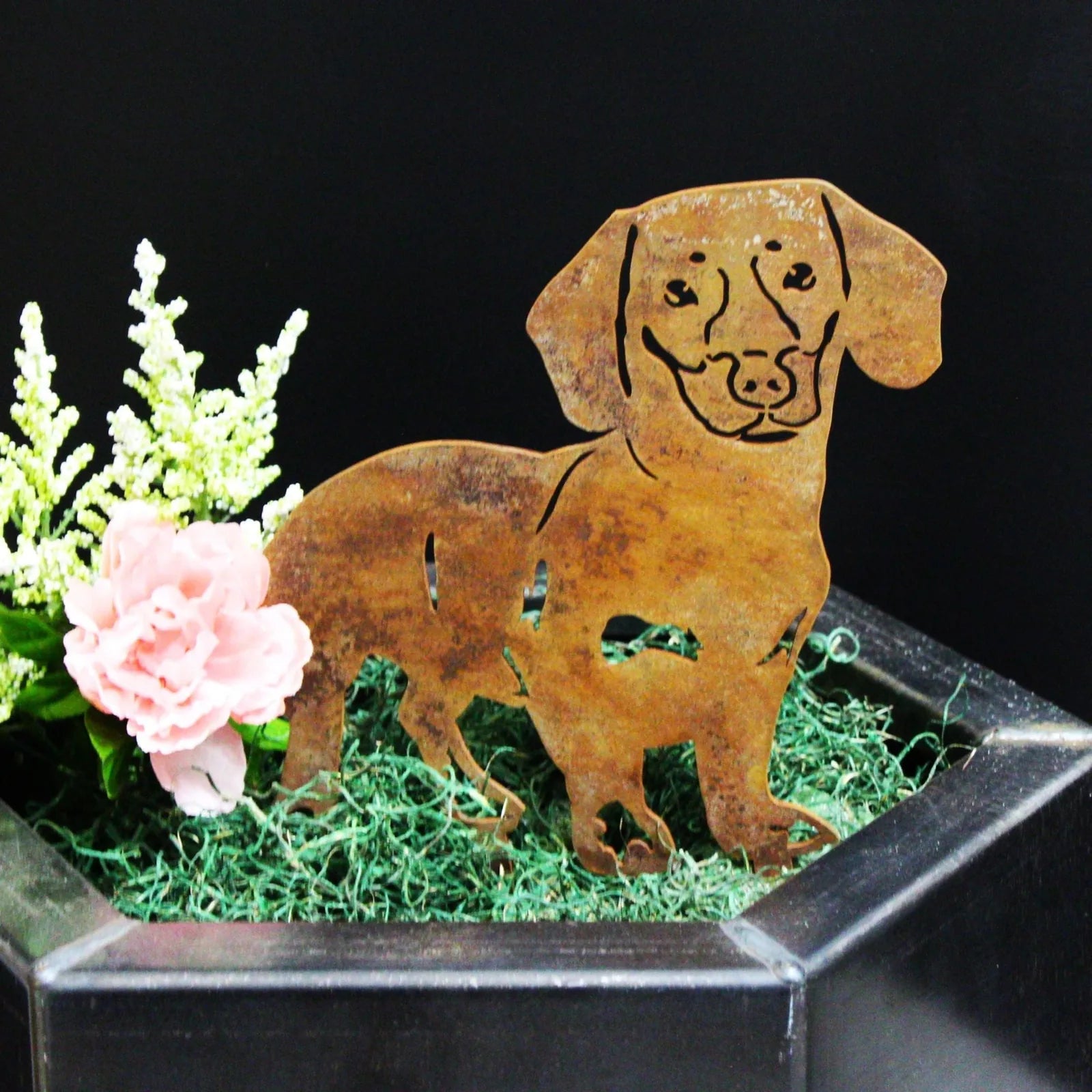 Corten steel silhouette of a happy Dachshund surrounded by green moss and a pink flower accent.