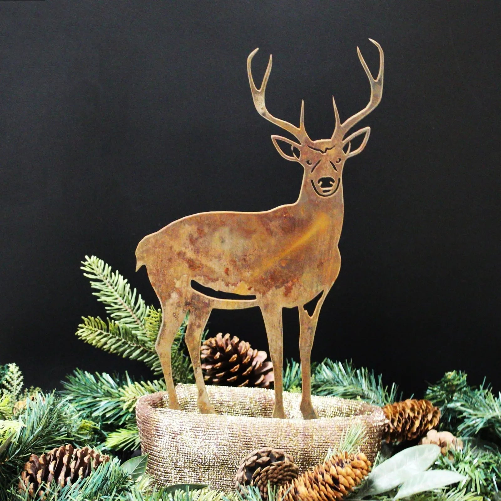 Rustic deer silhouette in weathered steel, surrounded by pinecones and greenery on a dark background.