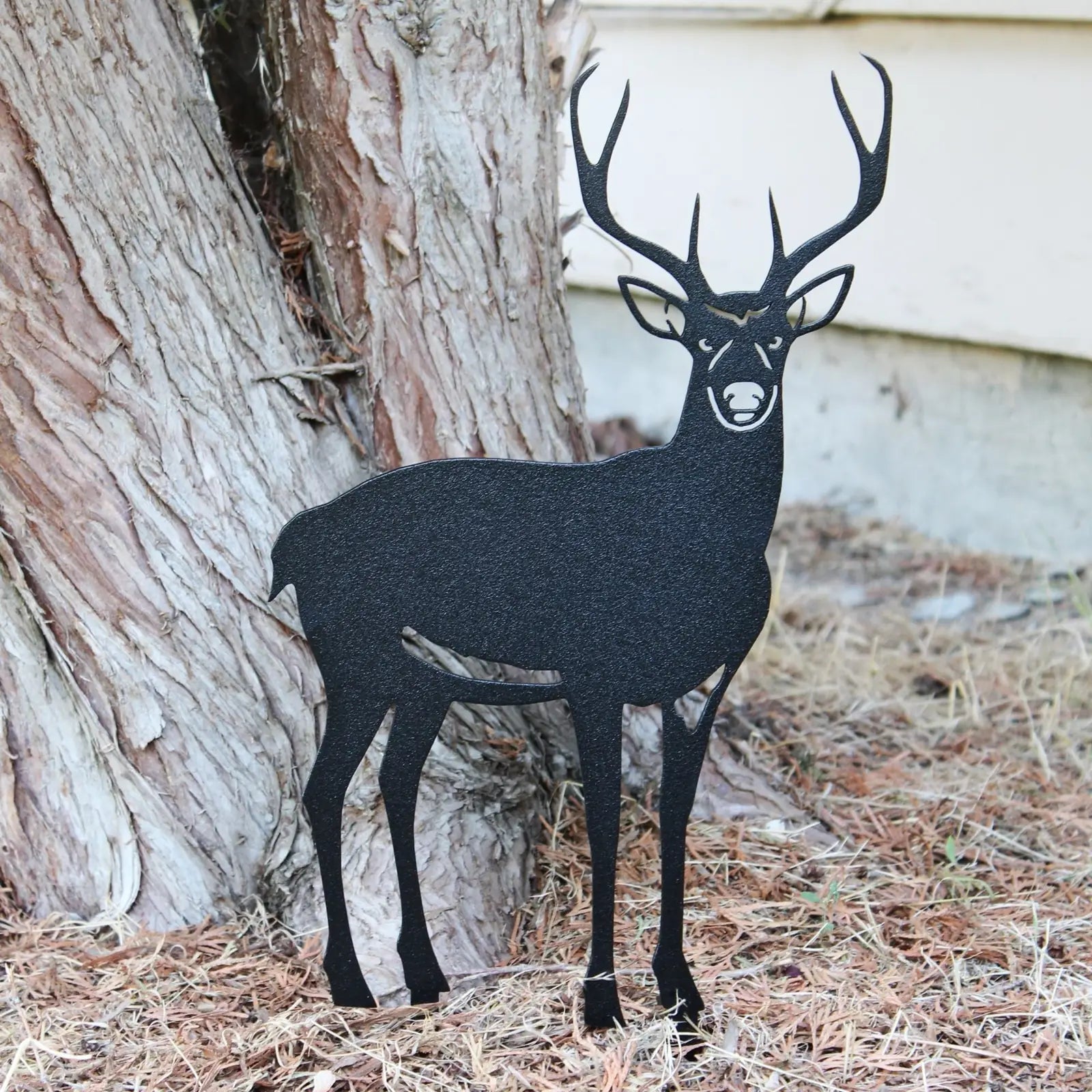 Black metal silhouette of a deer standing next to a tree, designed for outdoor decor in gardens or yards.
