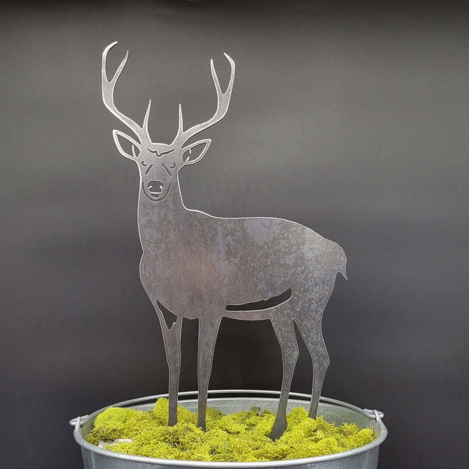 Metal deer silhouette in weathered Corten steel atop green moss, set against a dark background, perfect for outdoor decor.