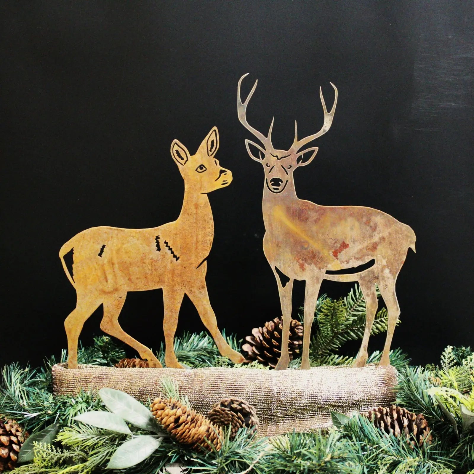 Rustic deer silhouettes made from Corten steel, surrounded by greenery and pine cones, on a dark background.