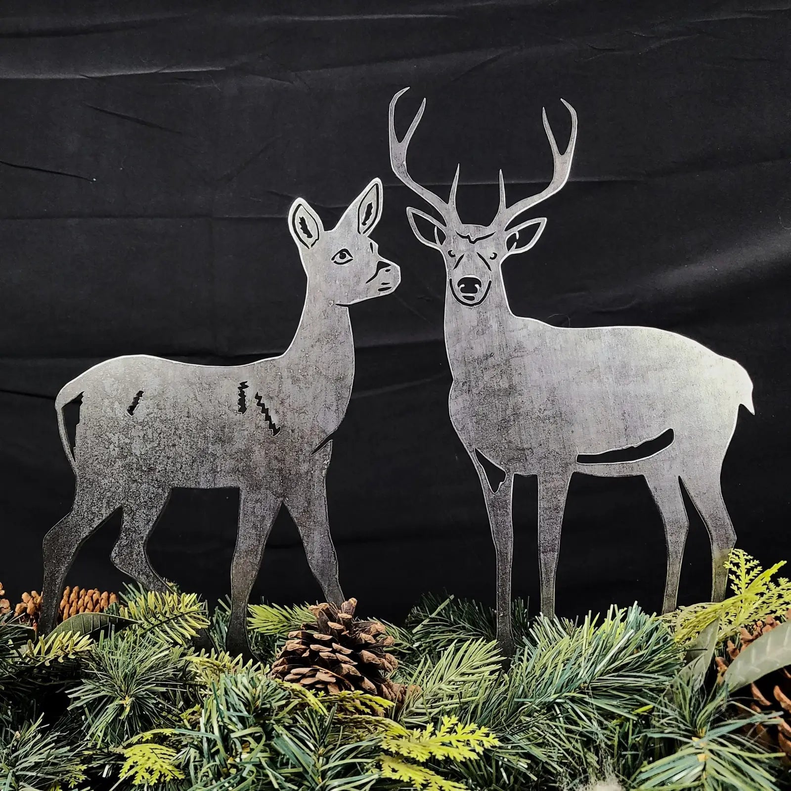 Handmade deer silhouette metal art in silver finish, depicting a doe and buck amid greenery and pinecones.