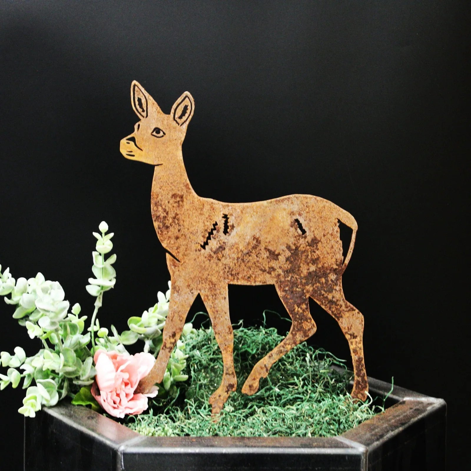Rusty deer silhouette art piece set against a black background, surrounded by greenery and a pink flower.