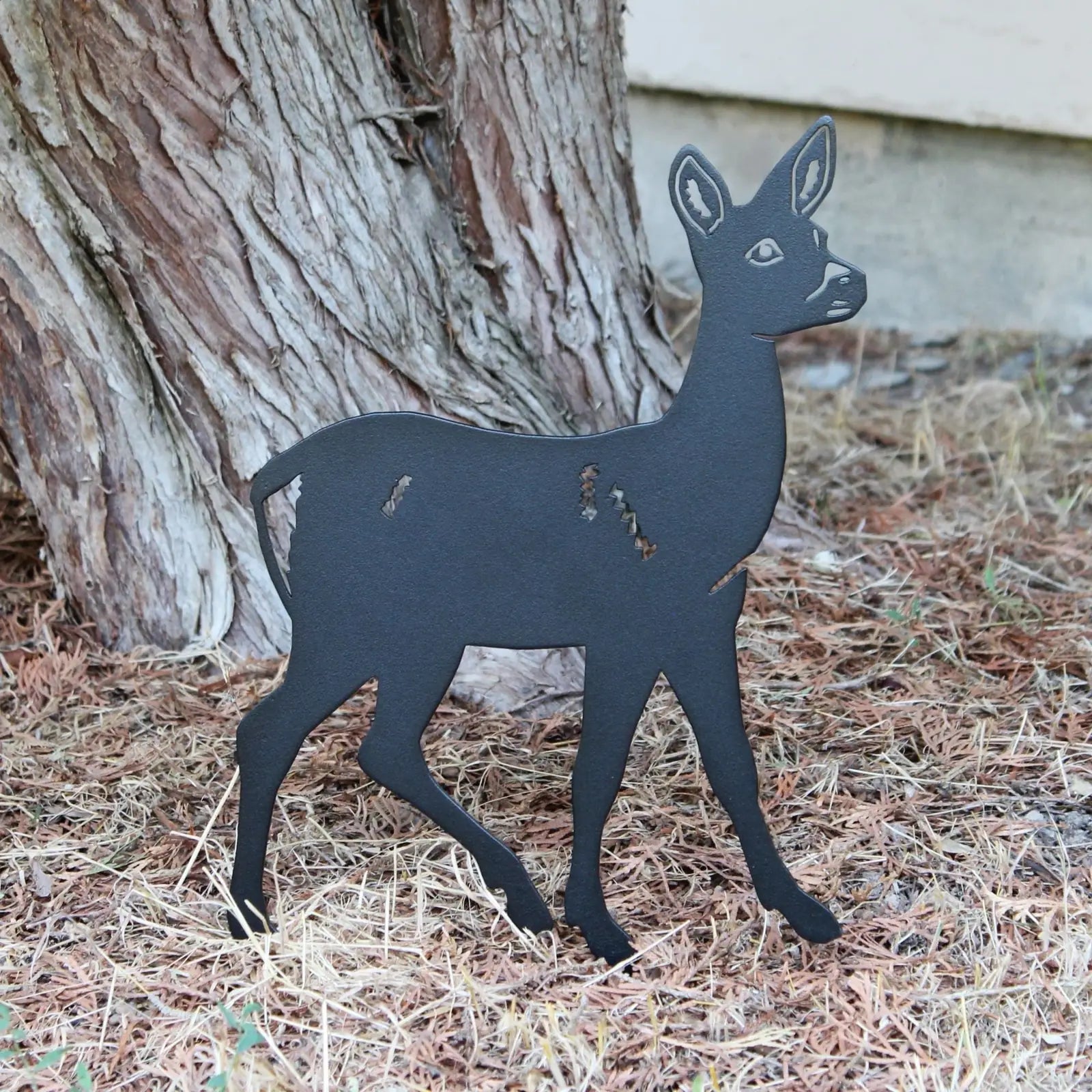 Black metal silhouette of a deer standing on dry grass near a tree, showcasing intricate cutouts and a sleek design.