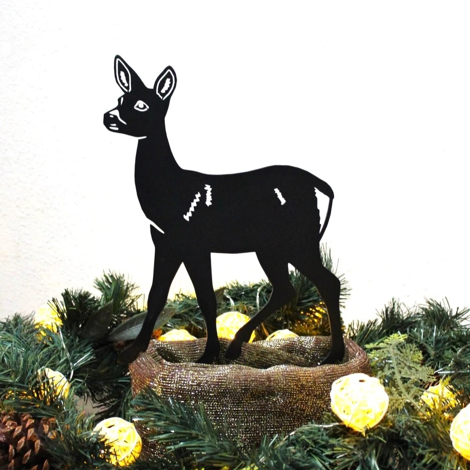 Stylish black deer silhouette made from metal, standing in festive greenery with soft glowing lights.