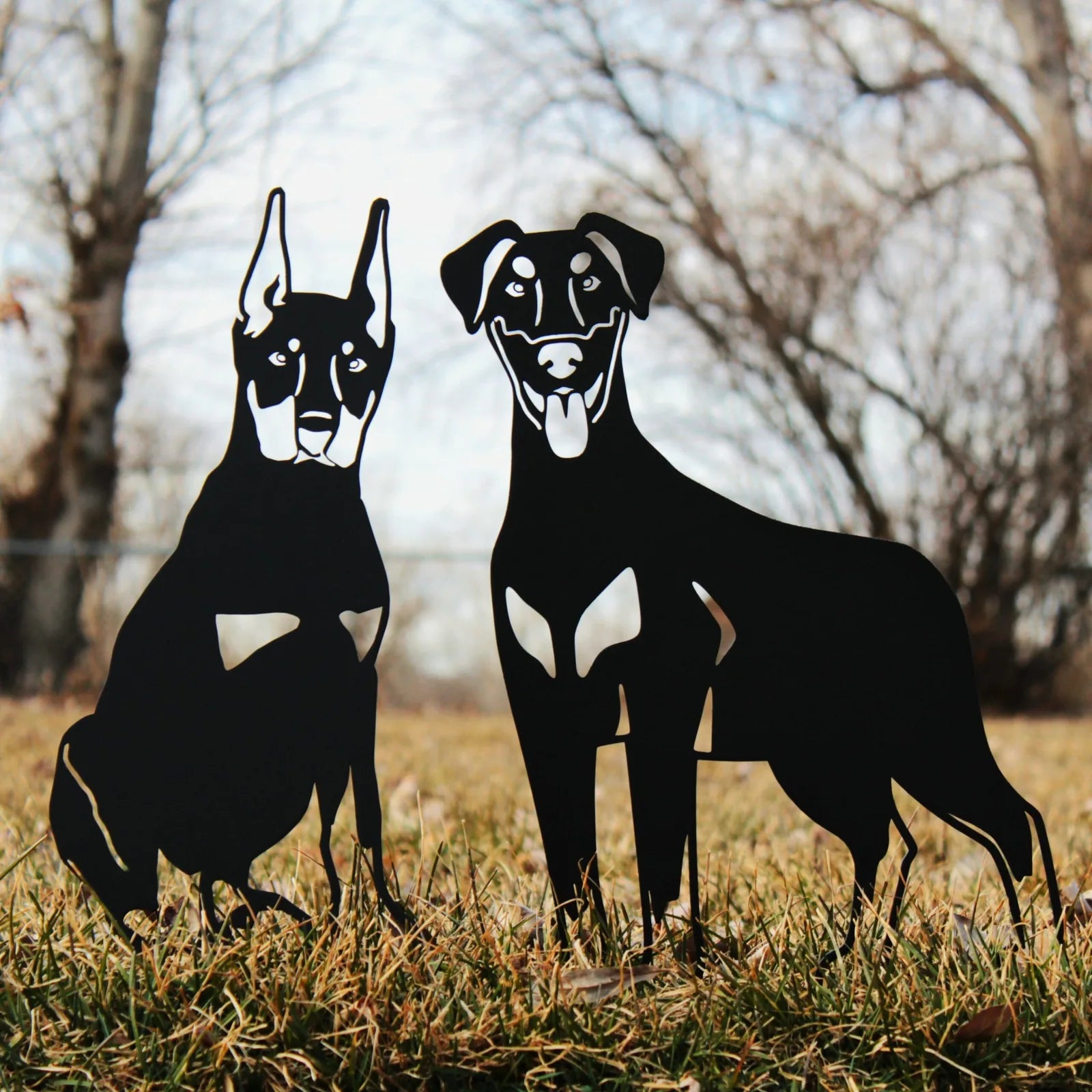 Black metal silhouette of a Doberman Pinscher and another dog, set on grass with a blurred background of trees.