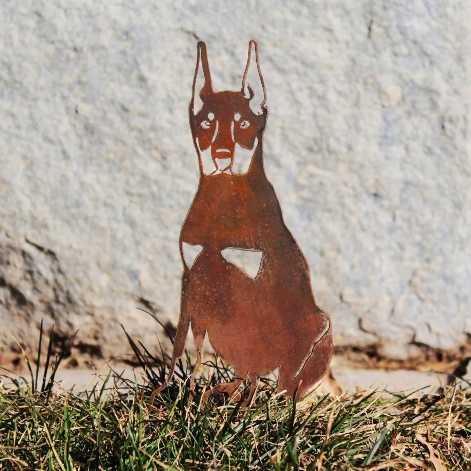Handmade Doberman Pinscher metal silhouette in weathered Corten steel, standing in grass against a stone backdrop.