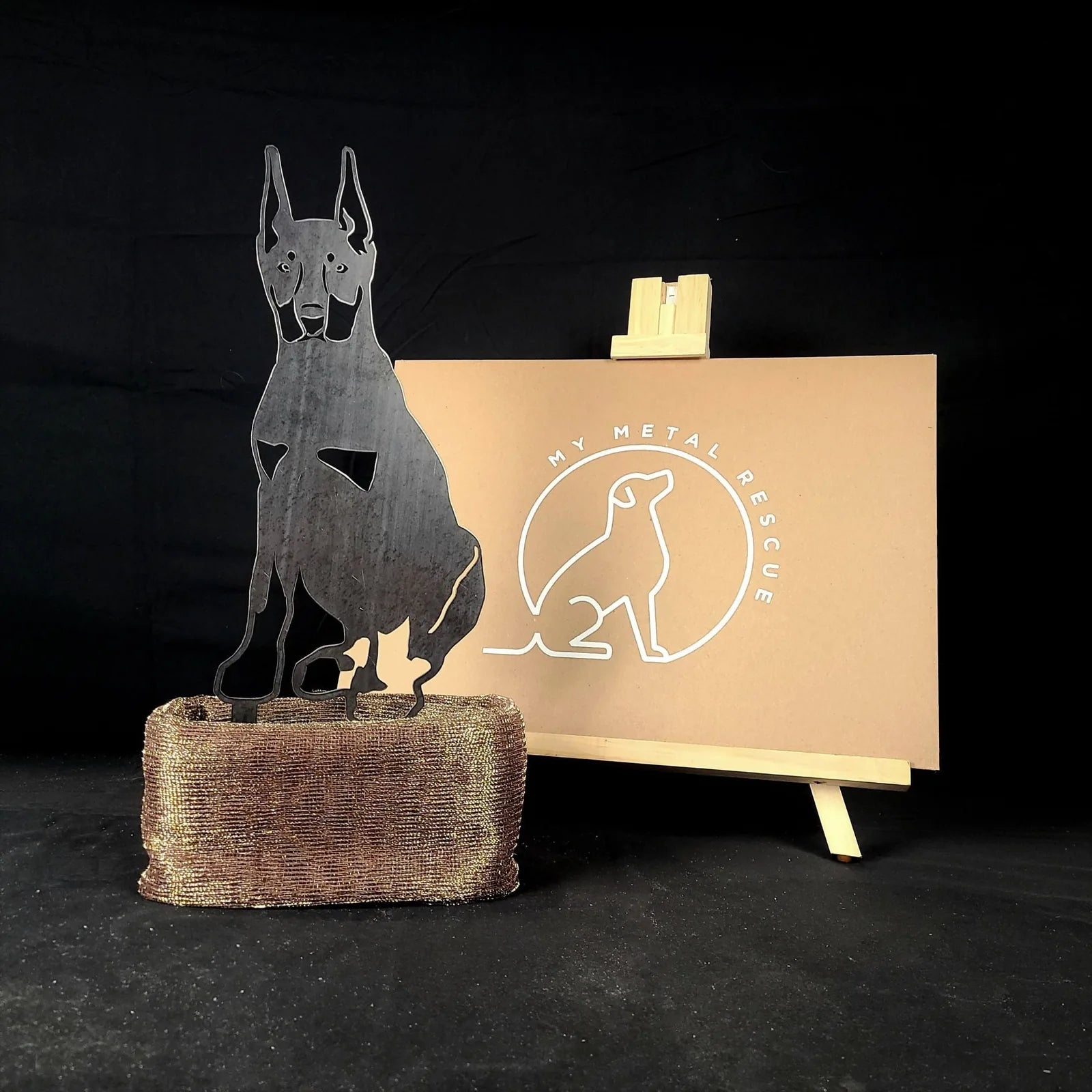 Cropped ear Doberman Pinscher metal silhouette in matte black, displayed on a textured base with branding backdrop.