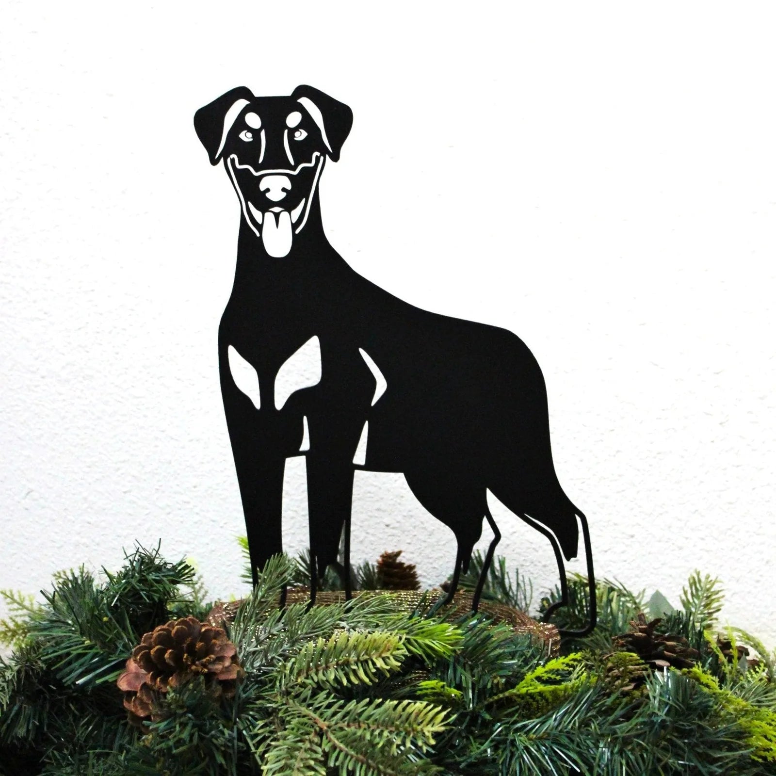 Black metal silhouette of a Doberman Pinscher with natural ears, placed on green foliage and pinecones for outdoor decor.