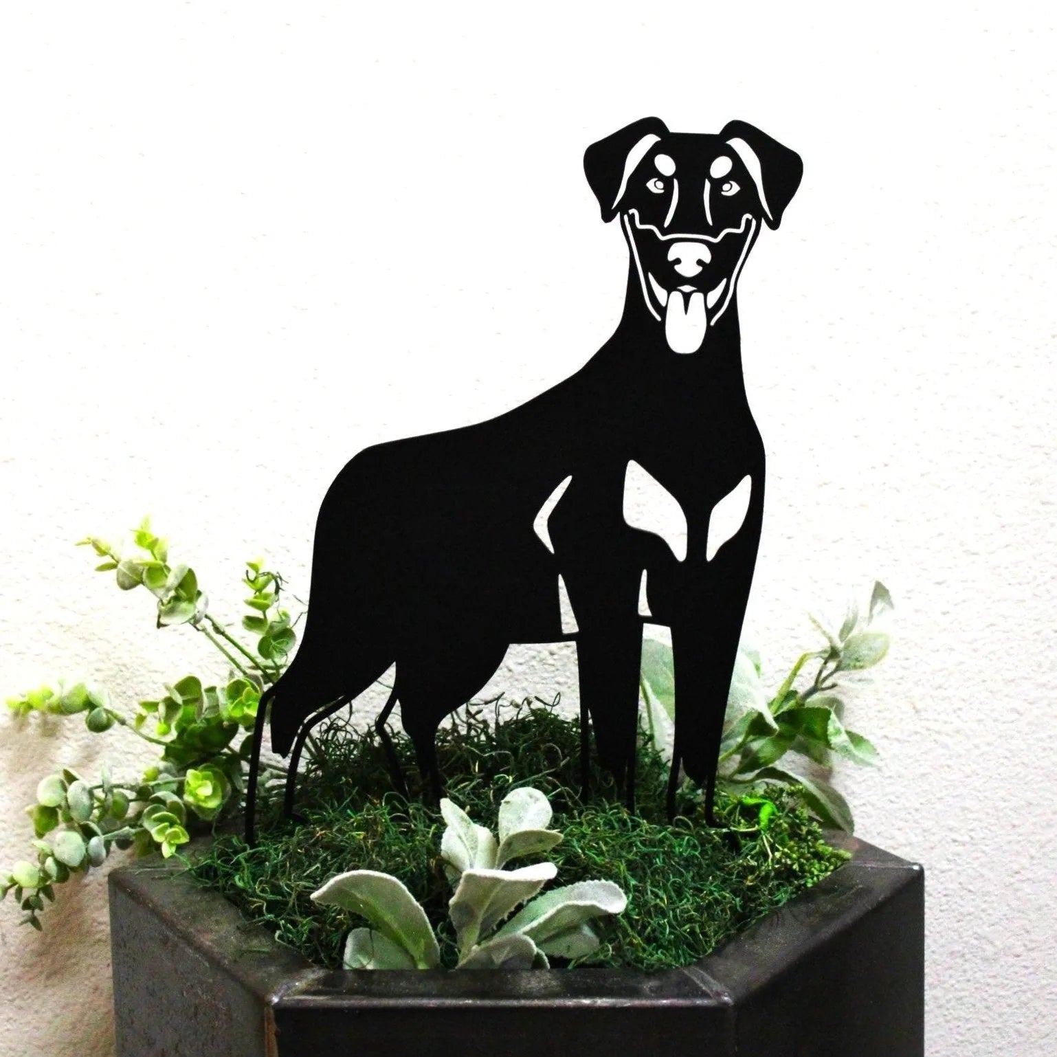 Black metal silhouette of a Doberman Pinscher standing in a planter with green foliage, showcasing outdoor dog decor.