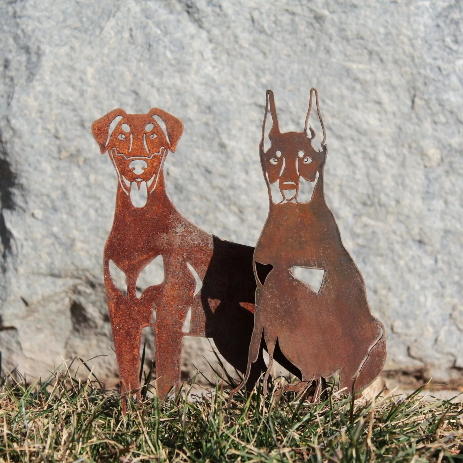 Rustic metal silhouettes of a Doberman Pinscher and another dog, showcasing intricate cut-outs, placed on grass.