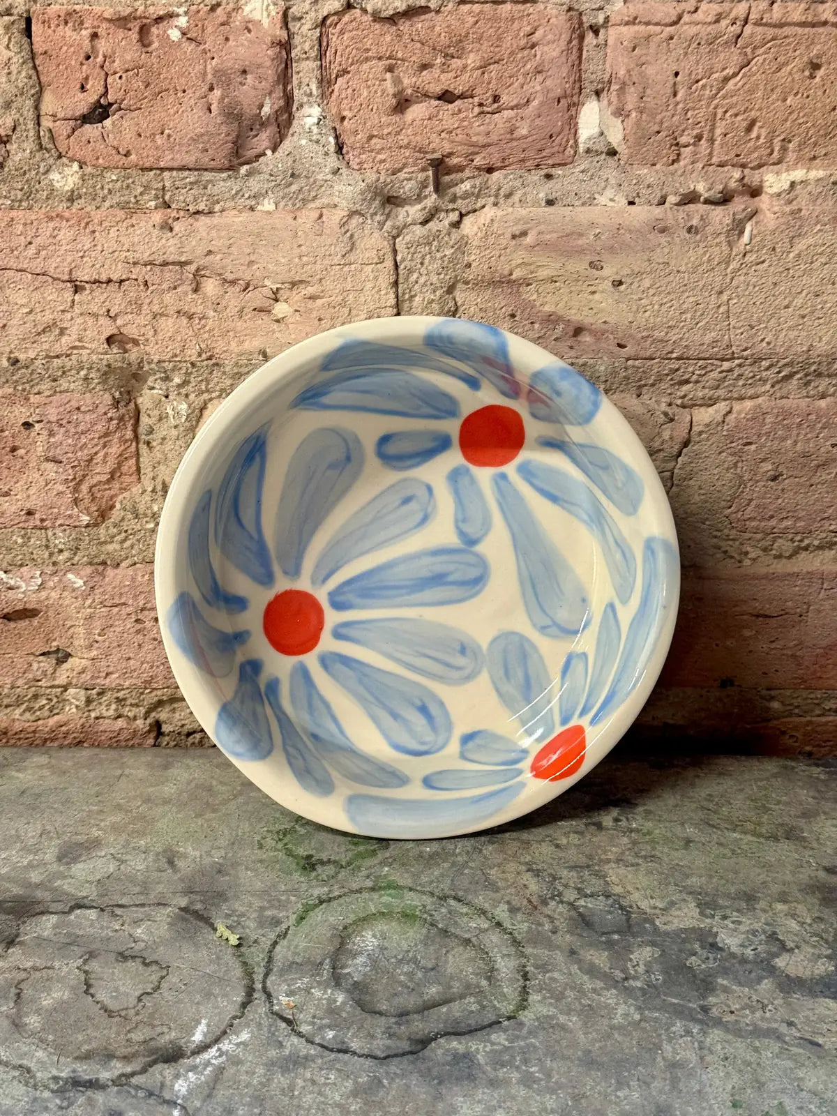 Hand-painted ceramic dog bowl featuring blue petals and orange centers, set against a rustic brick background.
