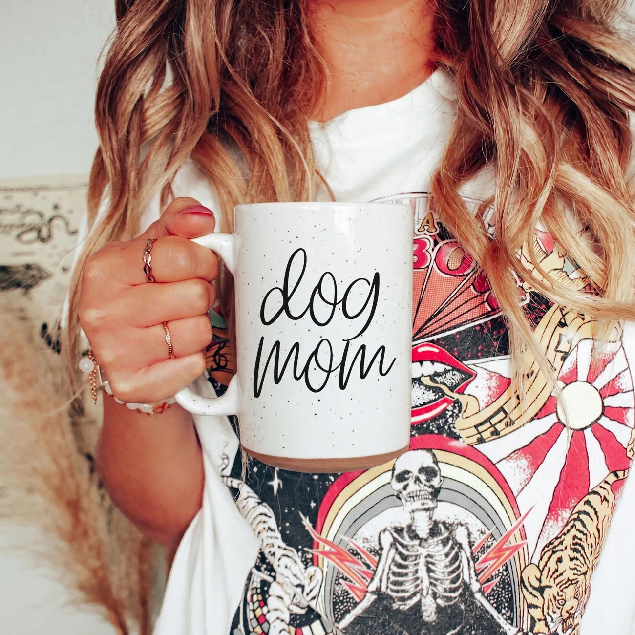 Off-white ceramic mug with dark speckles, featuring "dog mom" graphic in black, held by a person in a graphic tee.