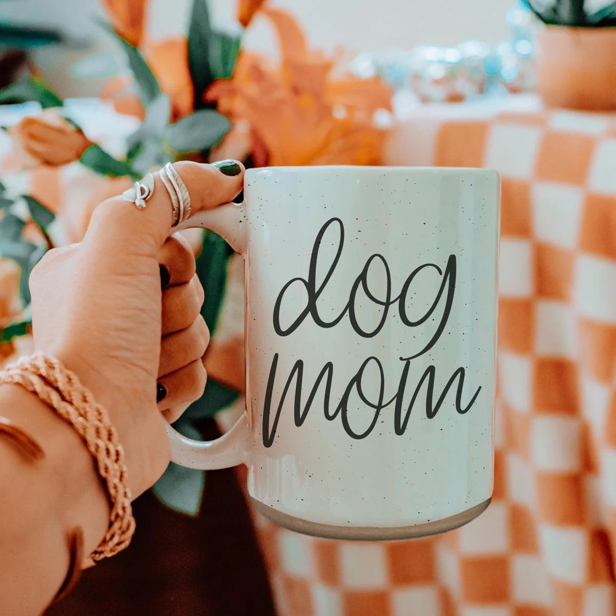 Off-white ceramic mug with dark speckles, featuring the text "dog mom," held against a floral background.