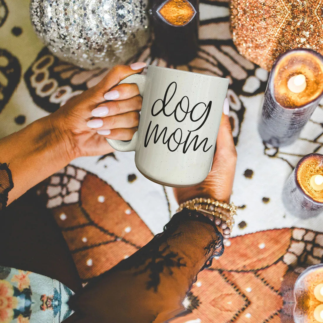 Off-white 16oz ceramic mug with "dog mom" graphic, held by a hand, surrounded by decorative candles and a patterned backdrop.