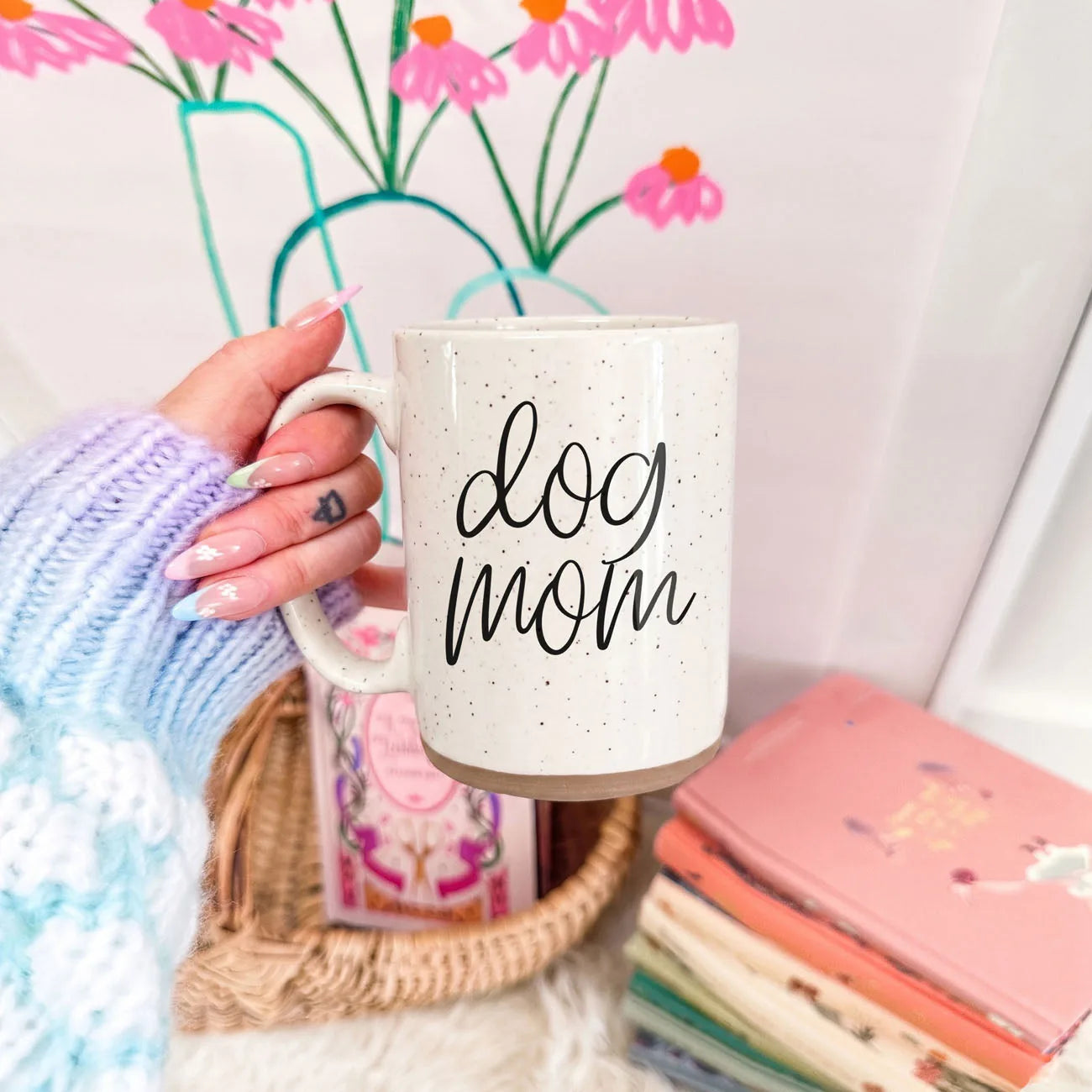 Off-white ceramic mug with speckles, featuring the "dog mom" graphic, held against a colorful floral backdrop.