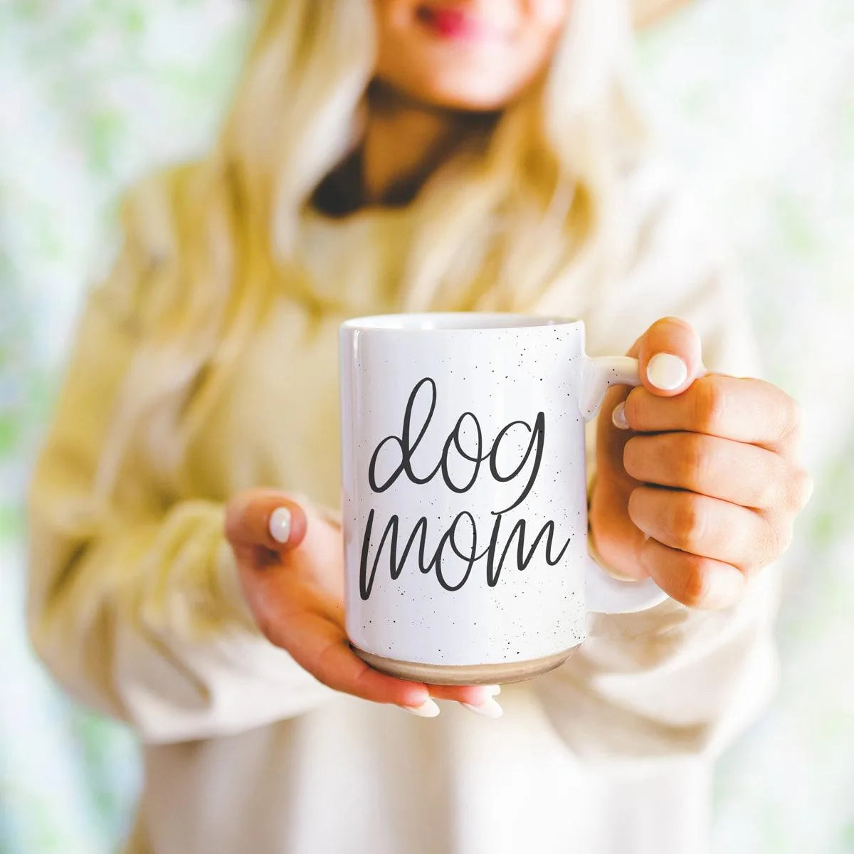 Off-white ceramic mug with dark speckles, featuring "dog mom" graphic; held by a woman in a cozy setting.