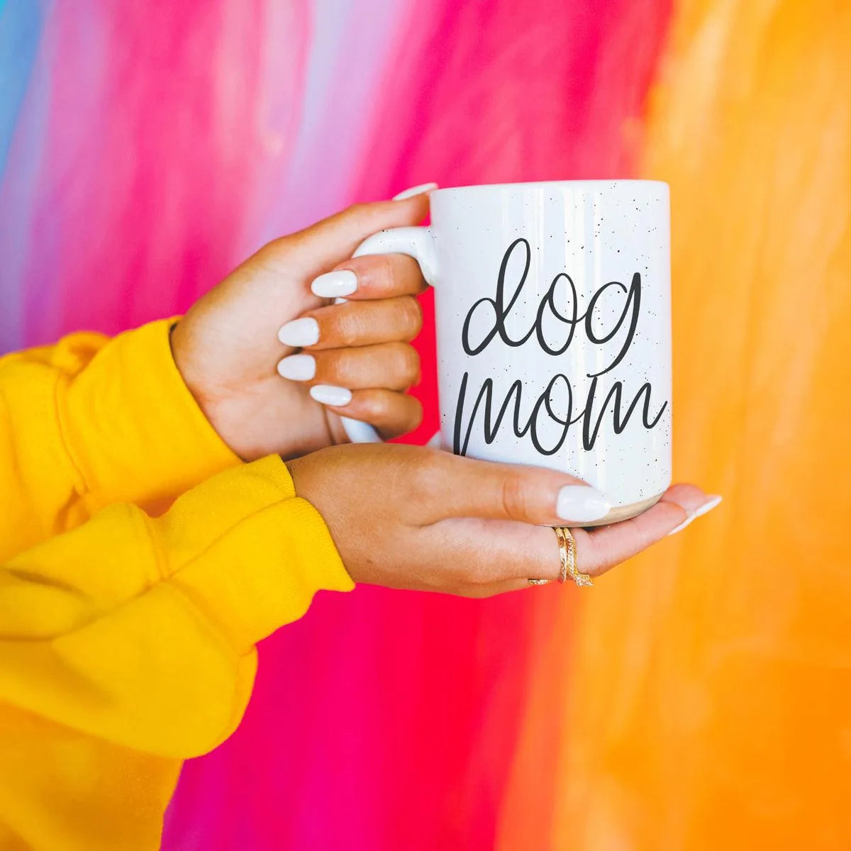 Off-white ceramic mug with black "dog mom" graphic, held by a person wearing a yellow sweater against a colorful background.
