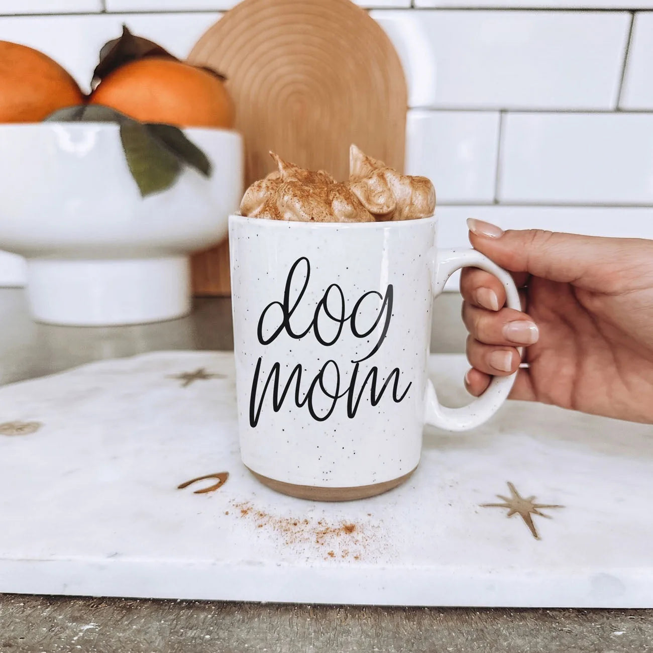 Handheld off-white ceramic mug with "dog mom" graphic, topped with whipped cream, set on a marble surface.