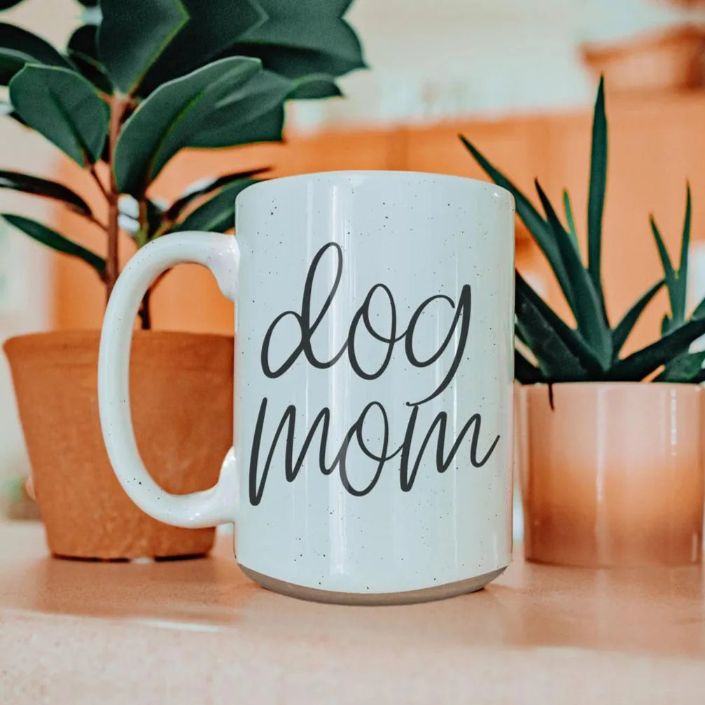 Off-white ceramic mug with dark speckles featuring "dog mom" graphic, surrounded by potted plants.