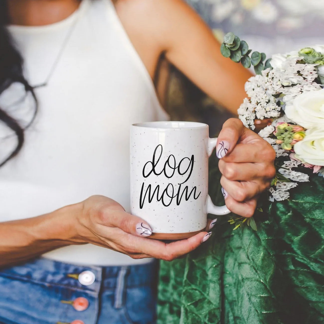 Off-white ceramic mug with black "dog mom" graphic, held by a person among greenery and flowers.