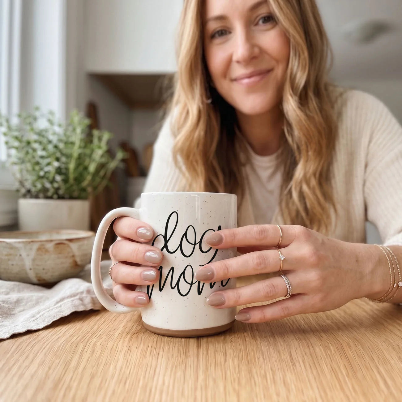 16oz off-white ceramic mug with "dog mom" graphic, held by a woman with light brown hair, sitting on a wooden table.