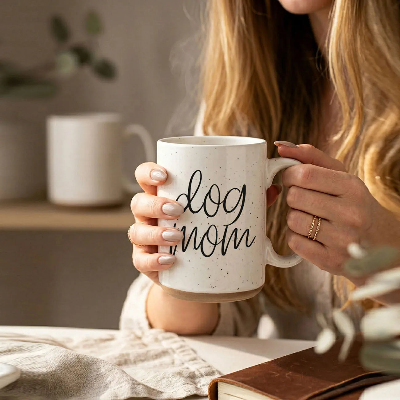 16oz ceramic mug with "dog mom" graphic, off-white with dark speckles, held by a person with long hair.