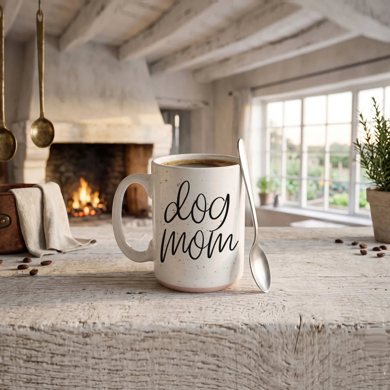 Off-white ceramic mug with "dog mom" text, speckled design, spoon beside it, set in a cozy, rustic room.