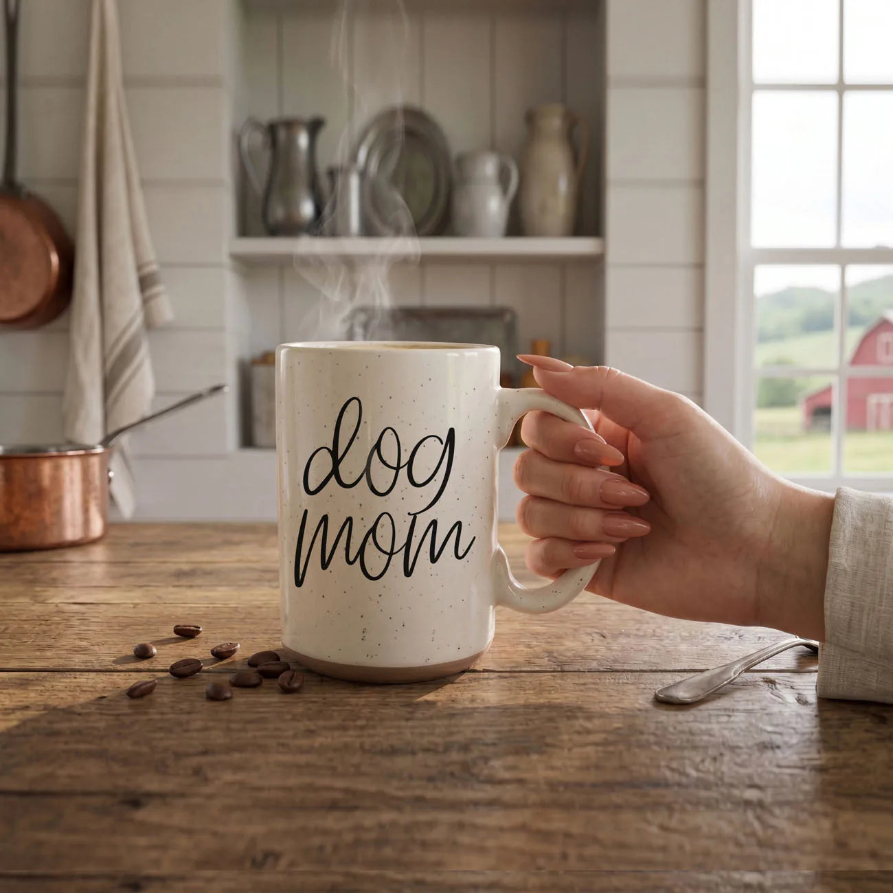 Off-white ceramic mug with dark speckles, featuring "dog mom" graphic, held in hand with steam rising, on wooden table.