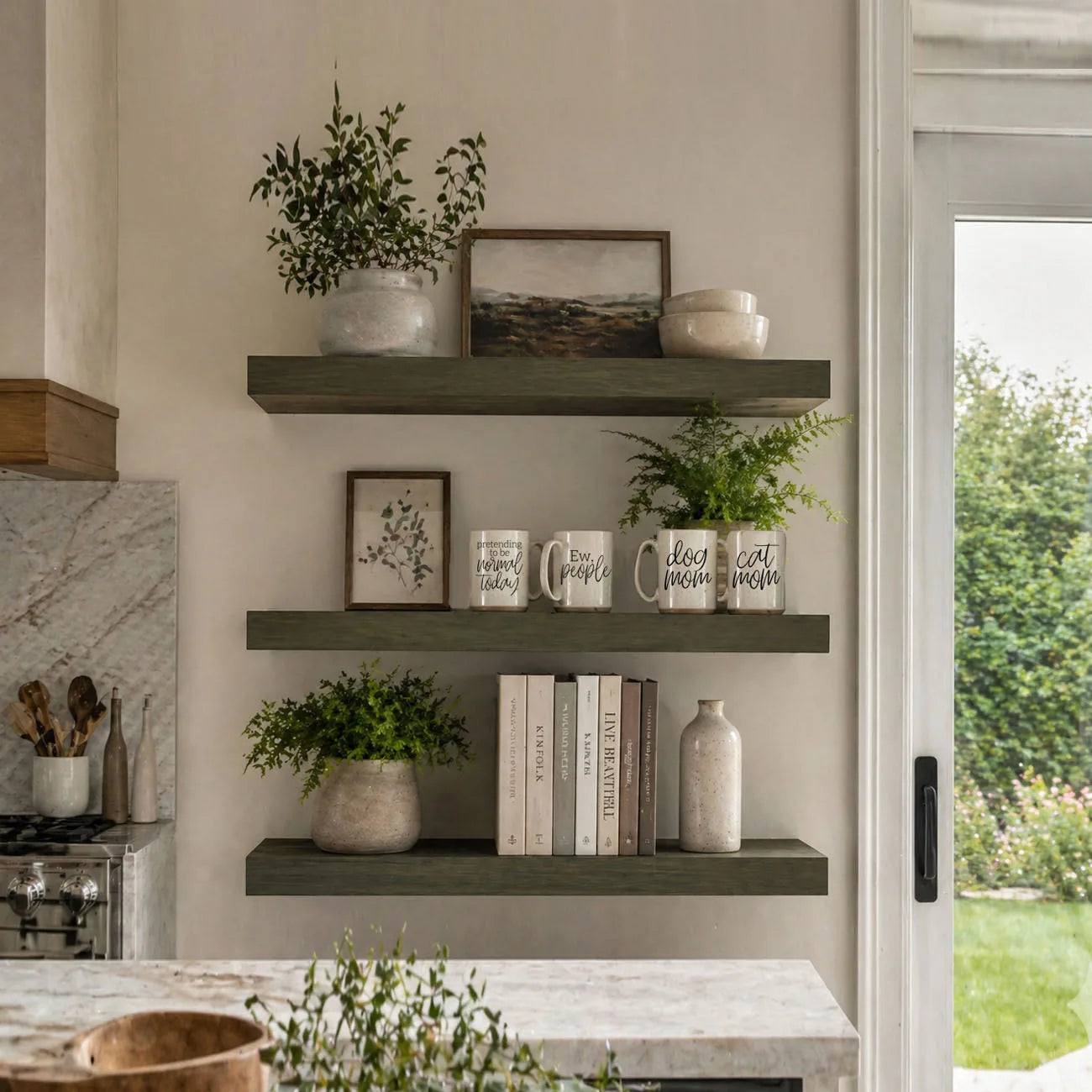 Ceramic coffee mugs on a shelf, including the "dog mom" mug, with green plants and books in a cozy kitchen setting.