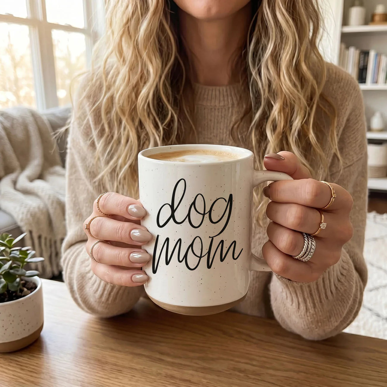 Cream-colored ceramic mug with "dog mom" graphic, held by a person wearing rings and cozy sweater.