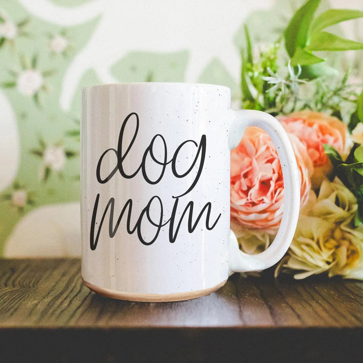 Off-white ceramic mug with a speckled design, featuring the "dog mom" graphic, surrounded by flowers.