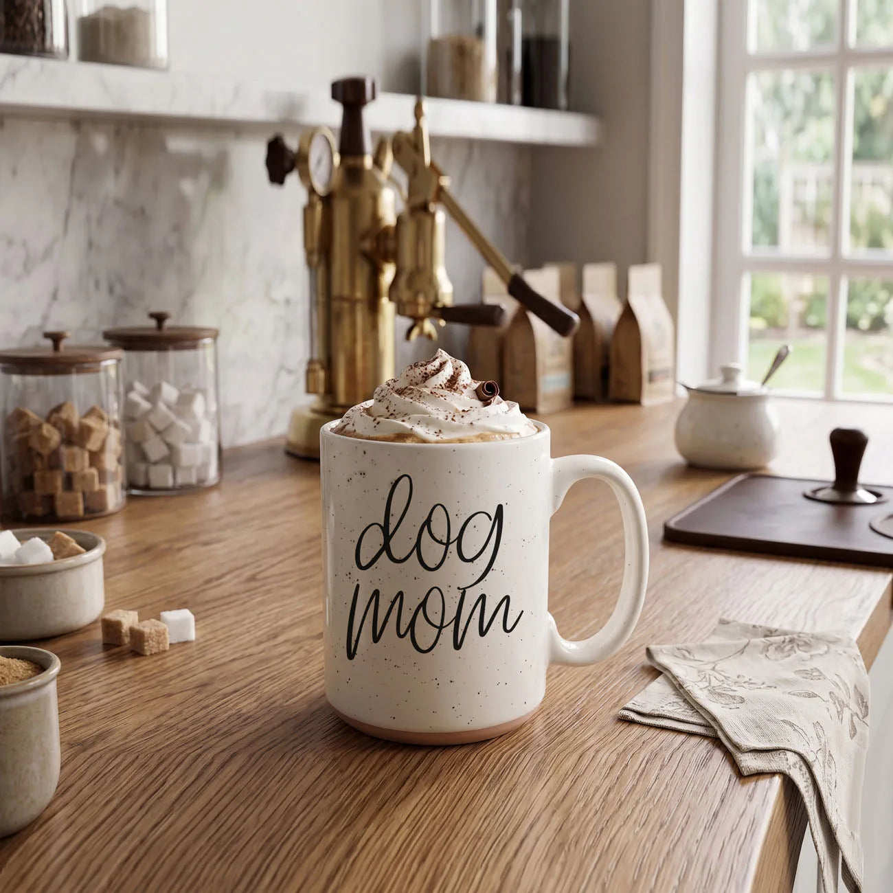 Off-white ceramic mug with dark speckles, featuring "dog mom" graphic, topped with whipped cream, set on wooden countertop.