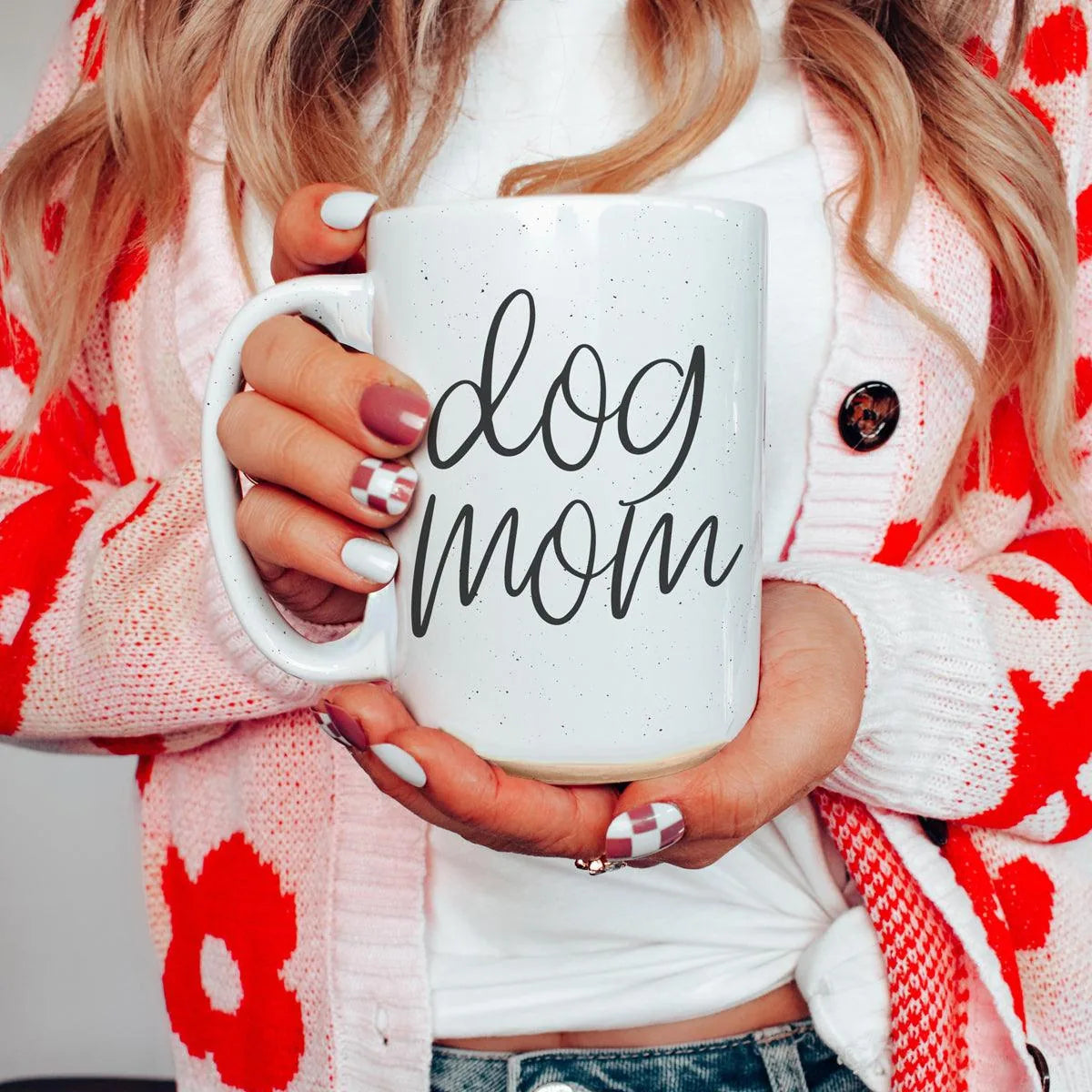 White ceramic mug with "dog mom" graphic held by a person in a red flower-patterned cardigan, showcasing colorful nail art.