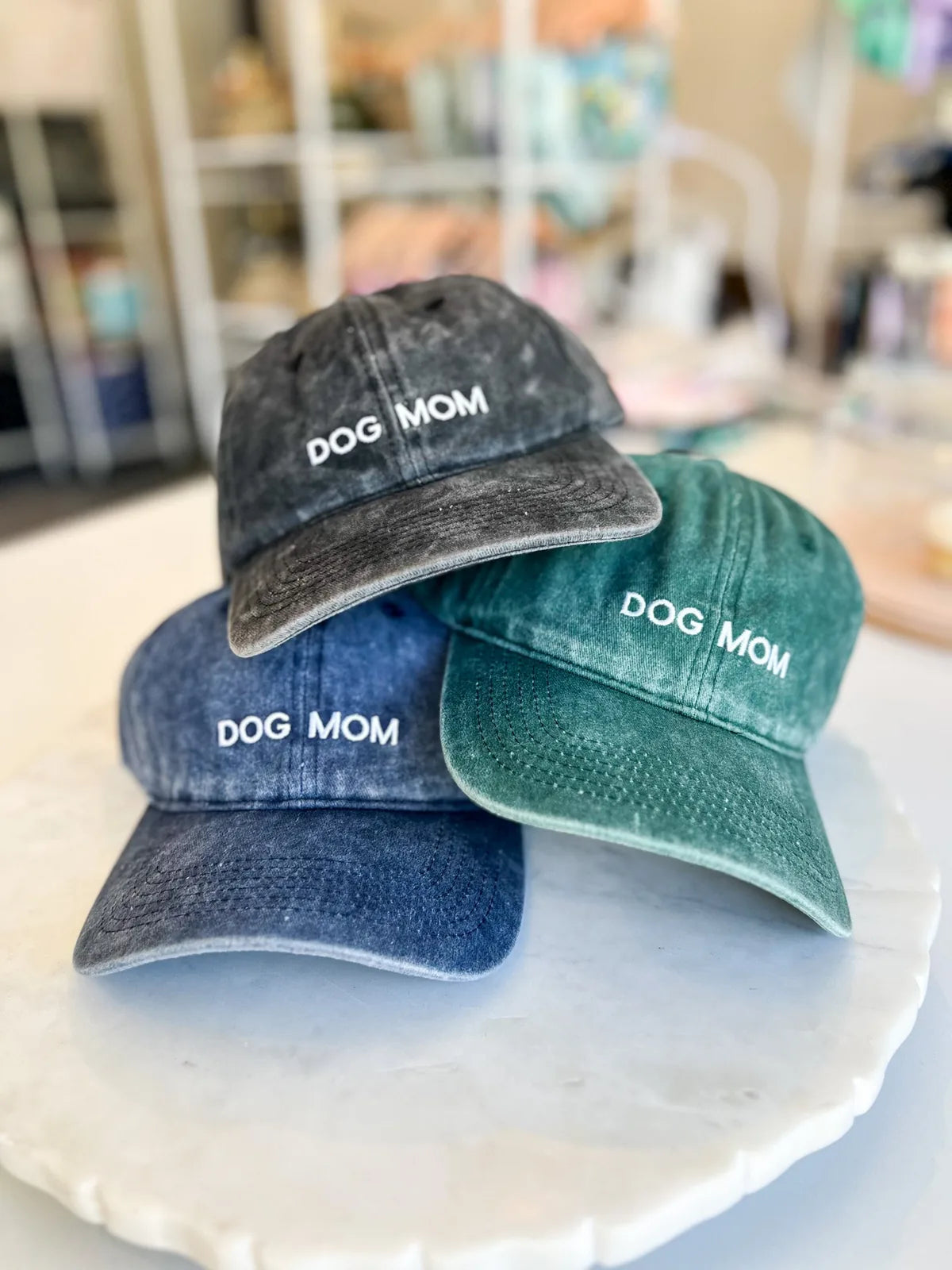 Three vintage-style dog mom hats in black, blue, and green on a marble surface, featuring white "DOG MOM" embroidery.
