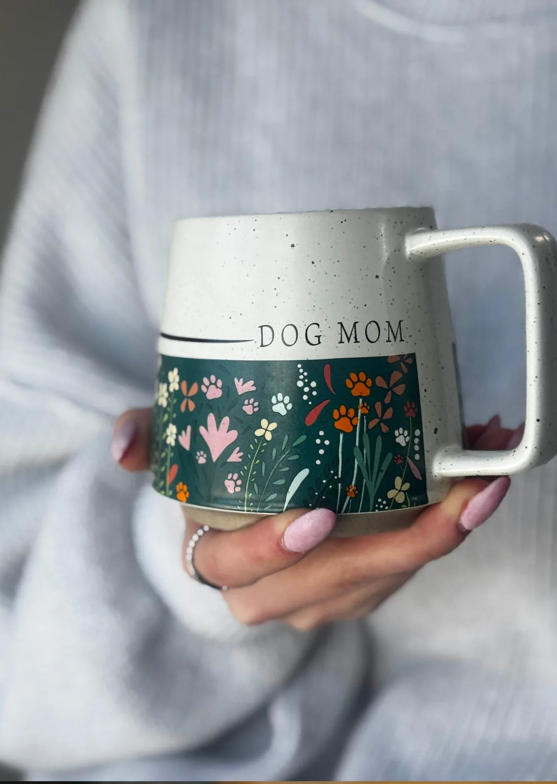 Ceramic Dog Mom mug featuring a dark floral design with hidden paw prints, held against a soft, textured sweater.