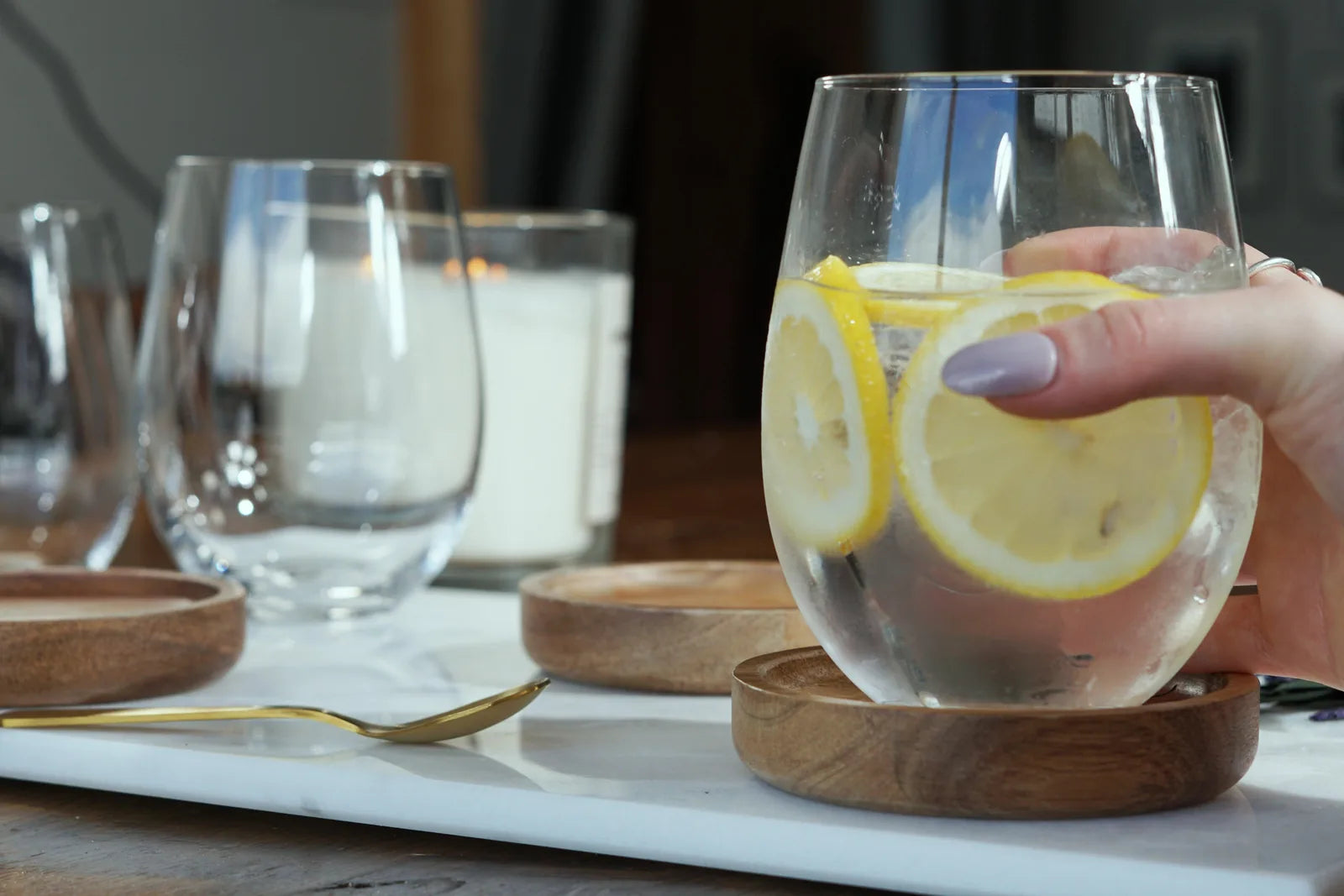 Glass filled with water and lemon slices, resting on a wooden coaster, with a hand holding it on a marble surface.