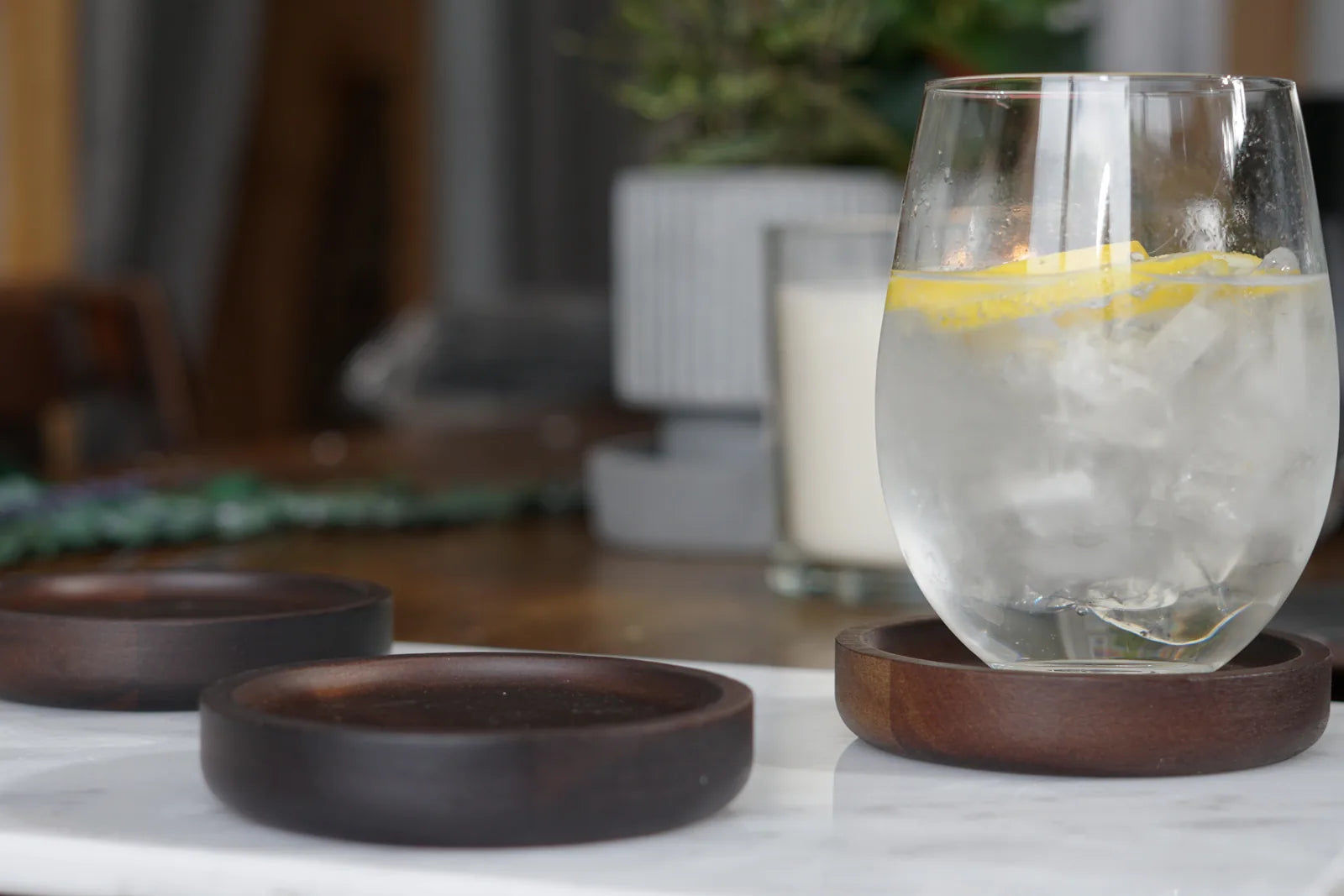 Clear glass with ice and lemon slice on a dark wood coaster, with two additional coasters on a marble surface.