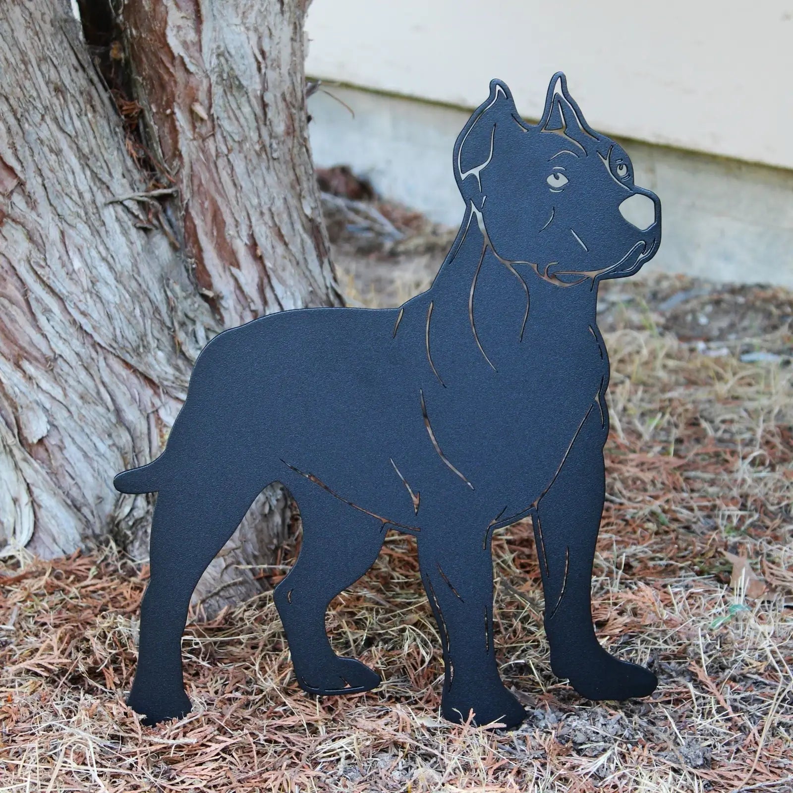 Dogo Argentino metal silhouette in matte black, standing among pine needles and tree bark.
