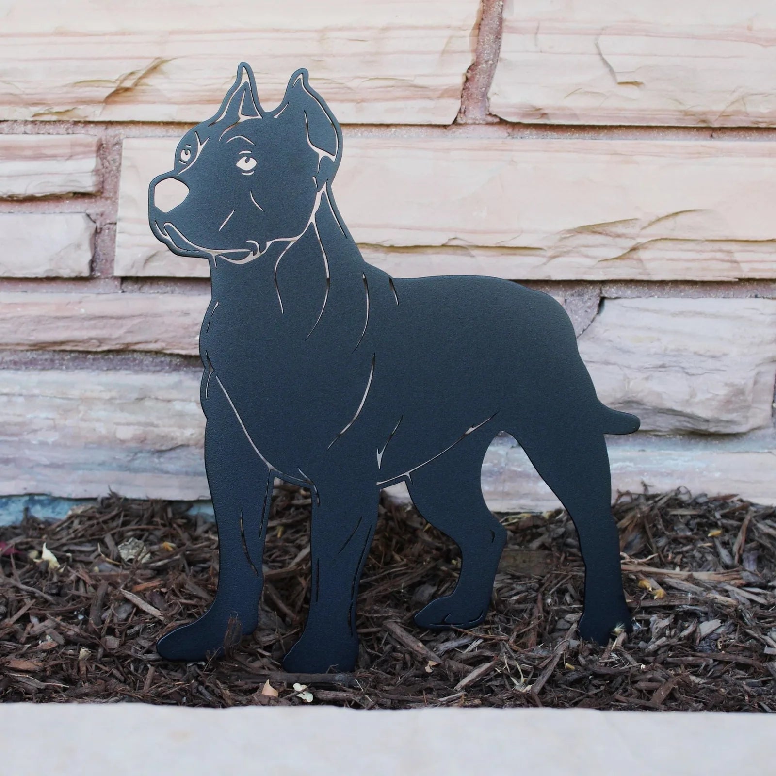 Dogo Argentino metal silhouette in matte black finish, standing on mulch against a stone wall backdrop.