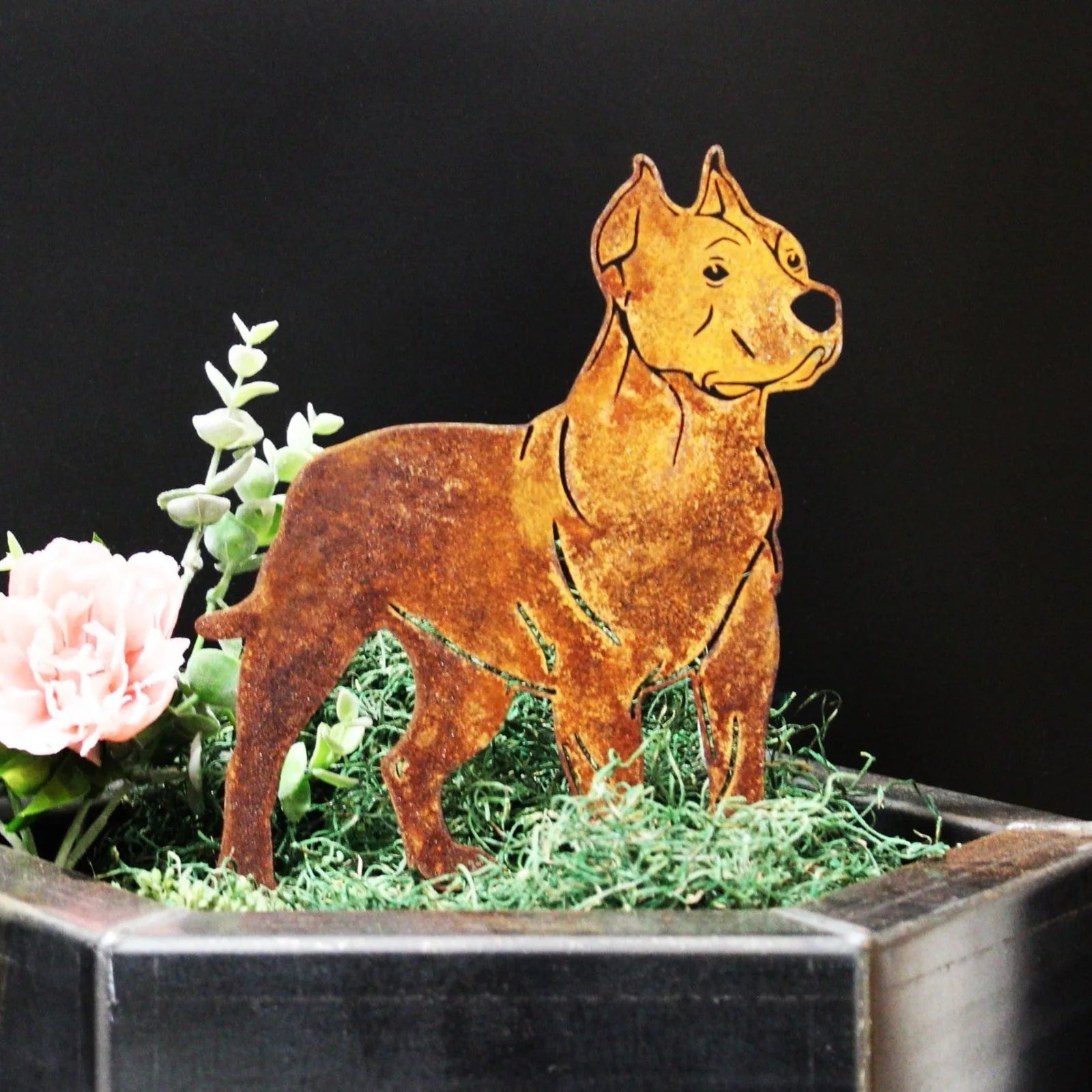 Dogo Argentino metal silhouette in weathered Corten steel, set among green moss and pink flowers in a decorative planter.