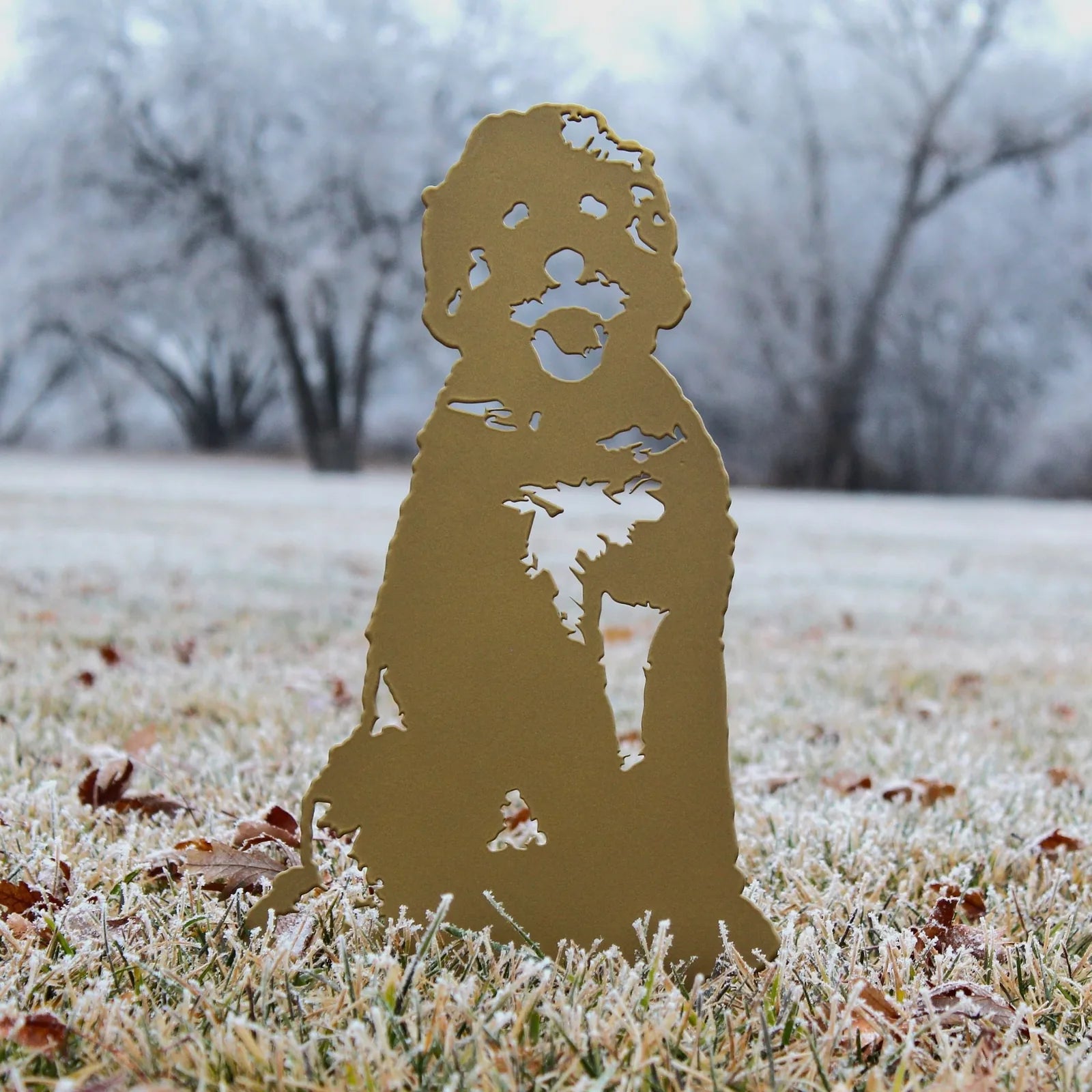 Gold Doodle metal silhouette displaying intricate details, set on frosty grass with trees blurred in the background.
