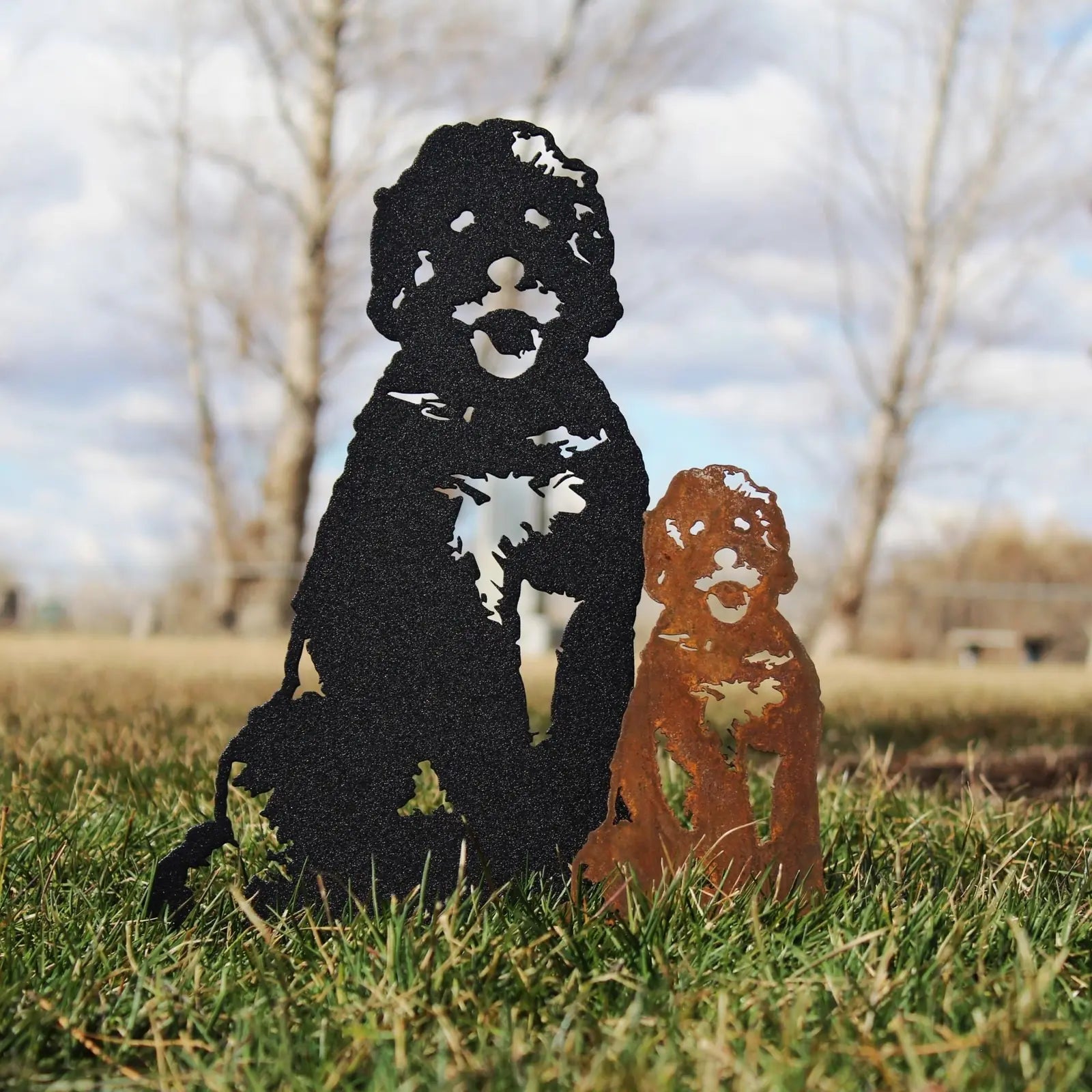 Black metal dog silhouette next to a rusted Corten steel puppy, set against a grassy outdoor background.