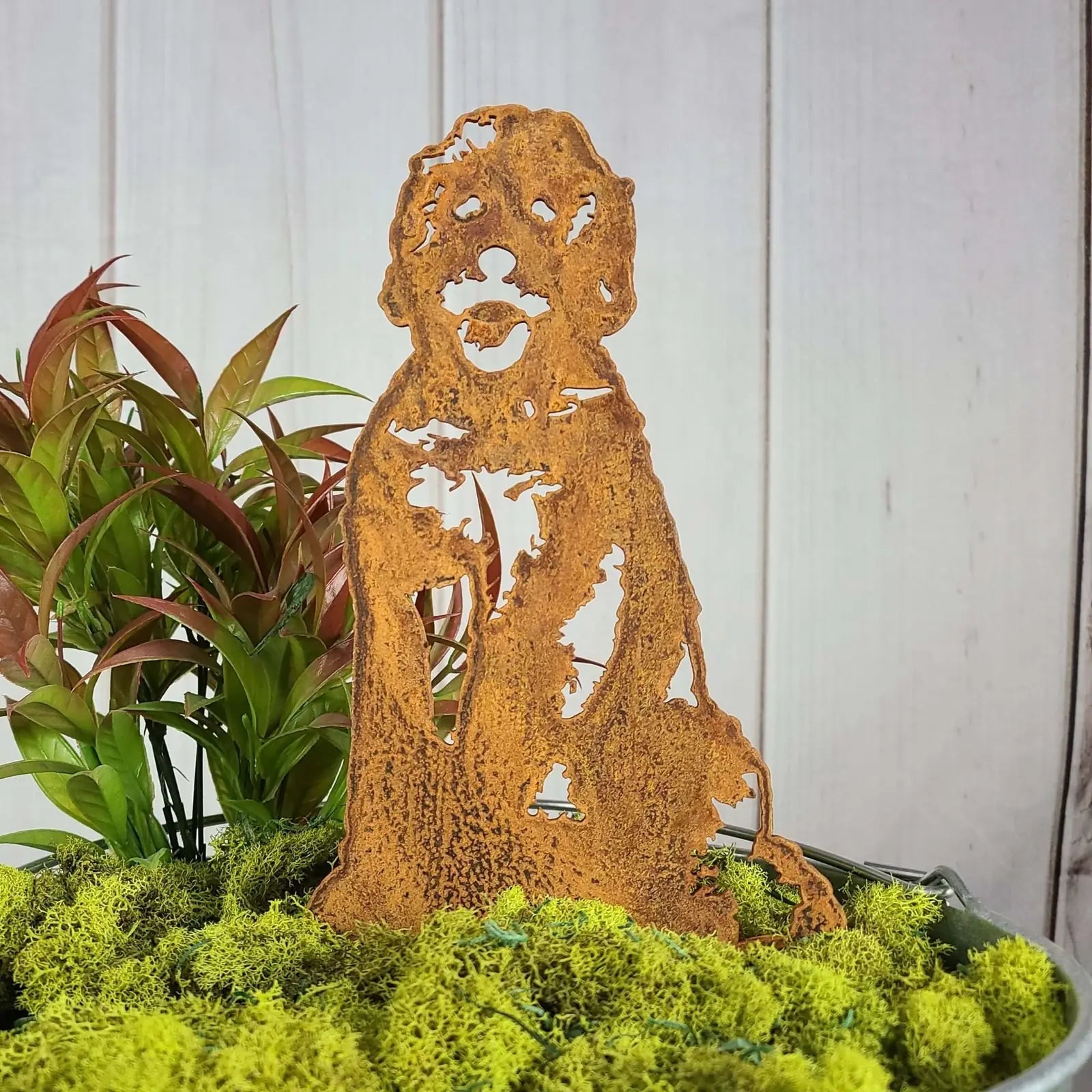 Rust patina Doodle metal silhouette surrounded by greenery and moss, adding charm to outdoor decor.