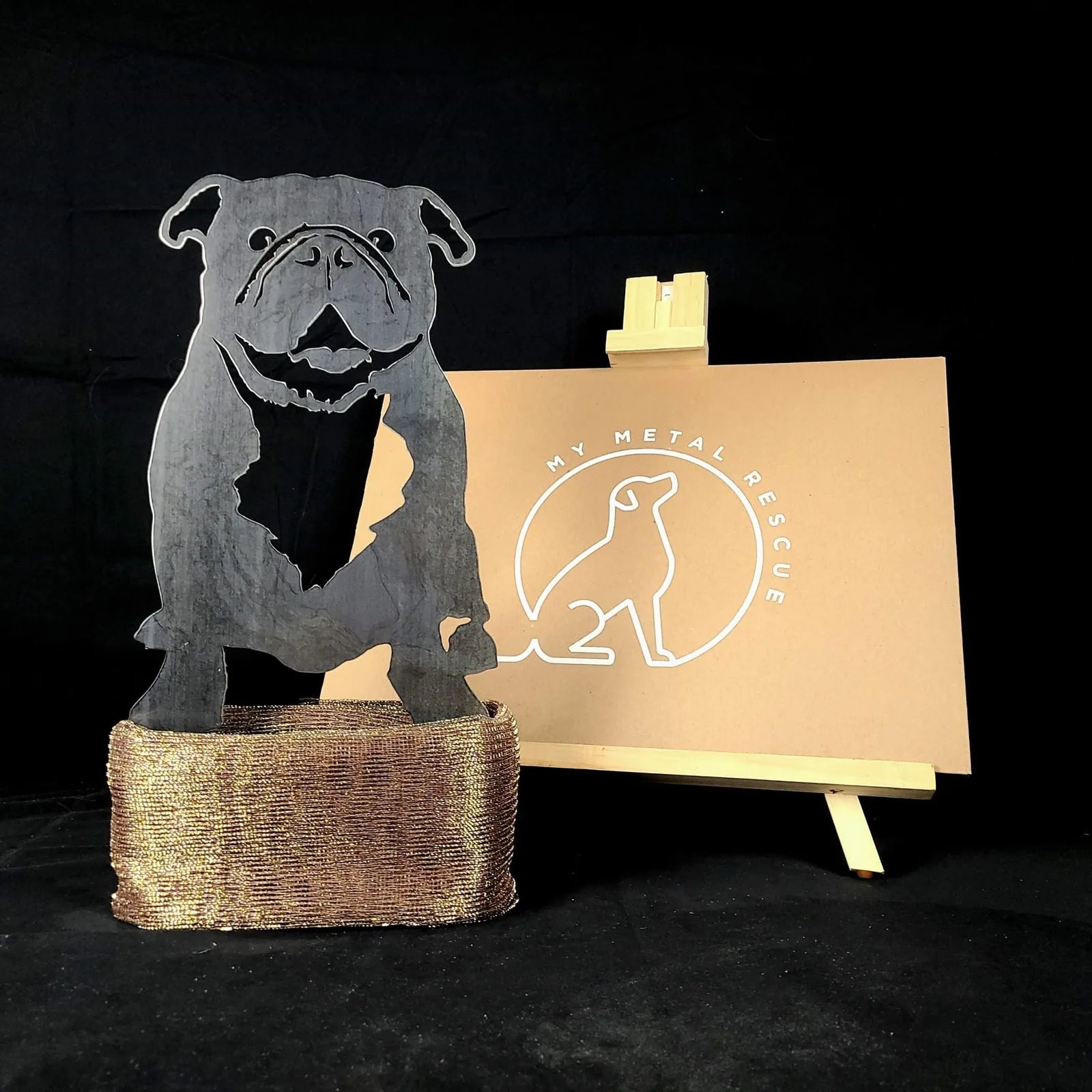 Black metal silhouette of an English Bulldog, displayed on a textured bronze base, with a logo in the background.