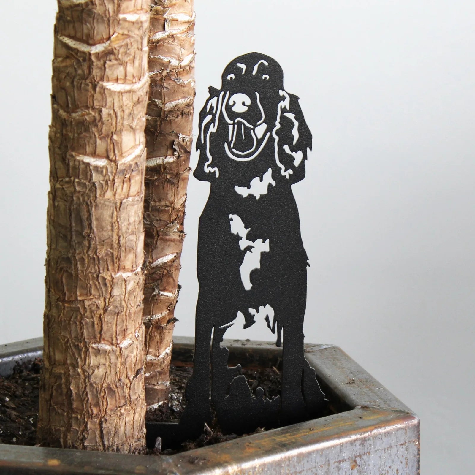 Black metal silhouette of an English Setter, placed beside a tree in a planter, showcasing outdoor dog art.