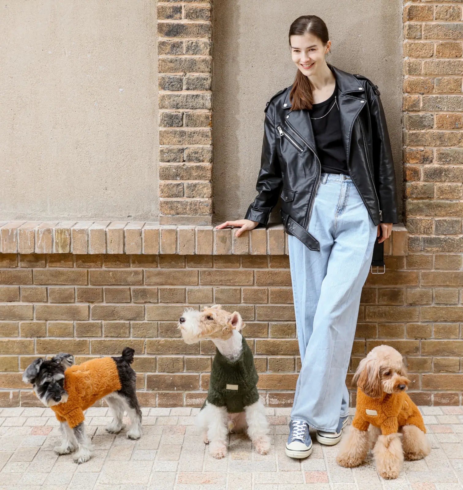 Three dogs wearing cozy sweaters, one in green and two in orange, standing beside a woman in a black leather jacket.